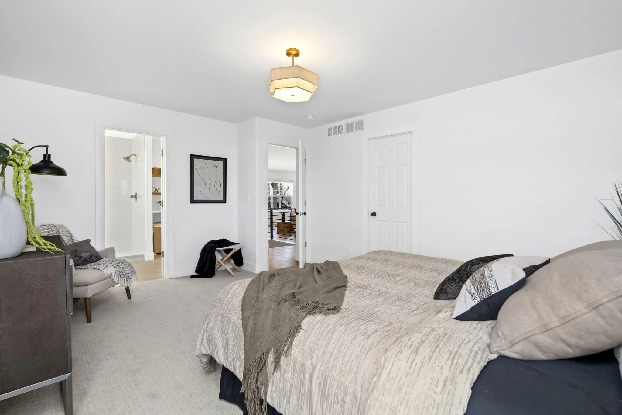 A bedroom with white walls, beige bedspread, black and white pillows, and a throw blanket. There is a small armchair with a throw, a black wall-mounted lamp, and a plant on a side table. Two doors, one leading to an en-suite bathroom and the other to