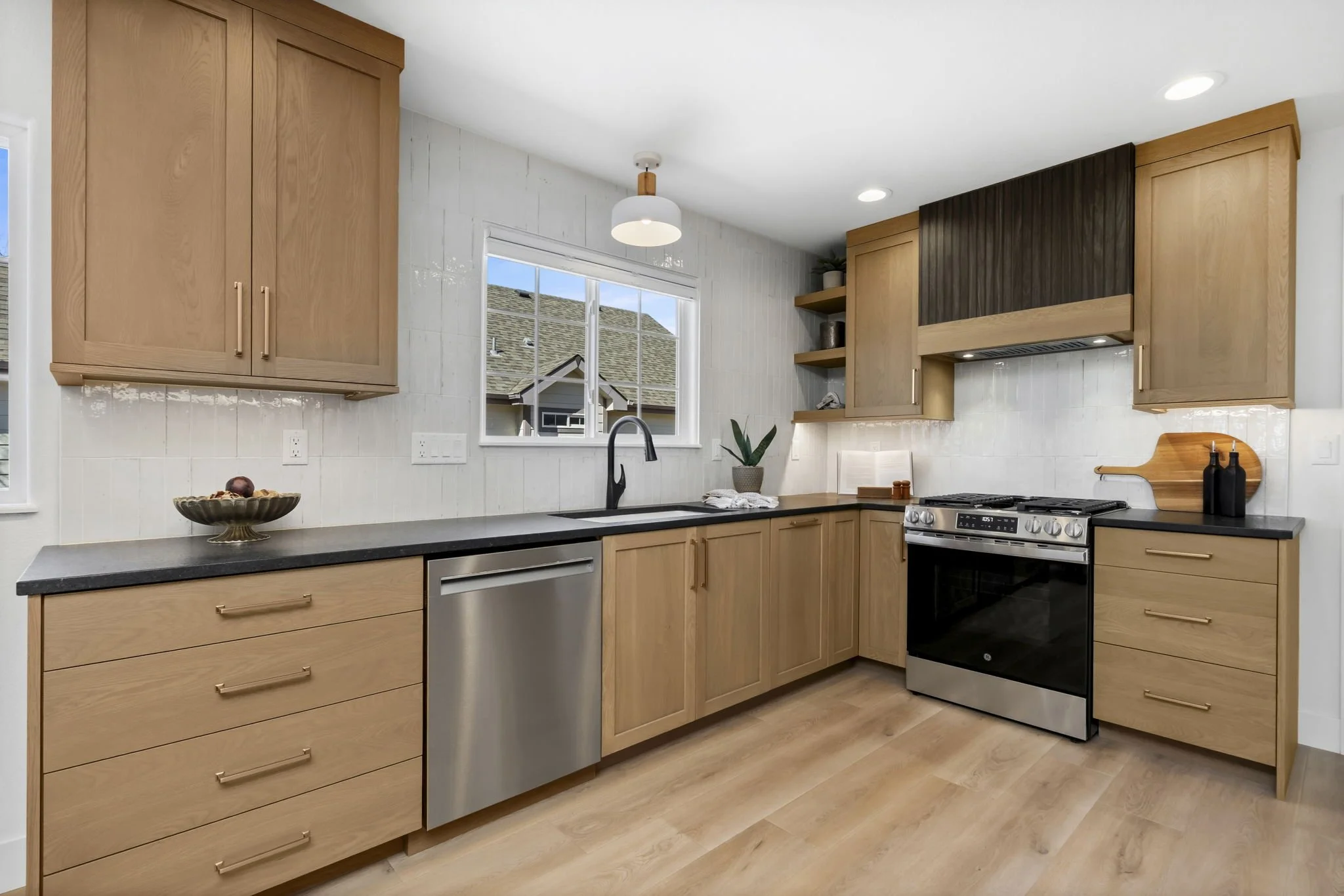 Modern kitchen with light wood cabinets, black countertops, a window above the sink, stainless steel dishwasher and oven, open shelving with decorative items, and a potted plant.