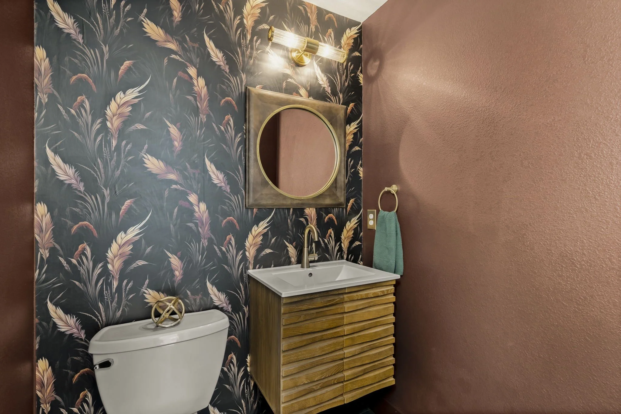 A bathroom with a dark floral wallpaper on the left wall, a square mirror with a gold frame above a small wooden vanity with a white sink and gold faucet, a wall-mounted light fixture with taillight bulbs above the mirror, a gold towel ring with a teal towel on the right wall, and a white toilet with a gold decorative item on top.