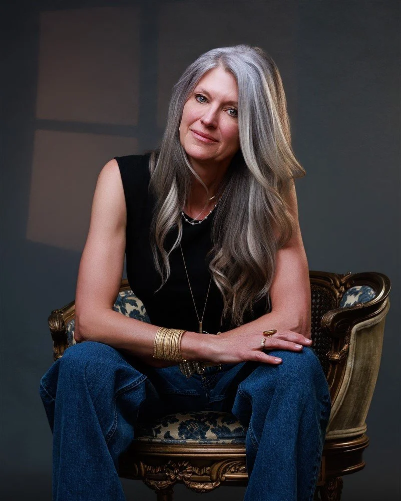 A woman with long gray hair sitting on an ornate vintage chair in front of a gray background.