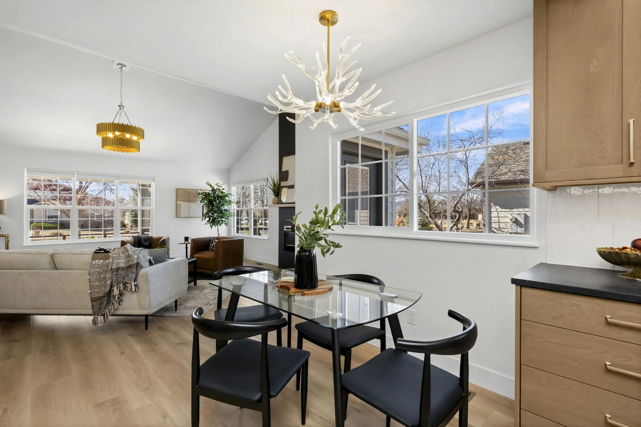 Open-concept living and dining area with large windows, modern chandeliers, beige sofa, brown armchairs, and a glass dining table with black chairs.