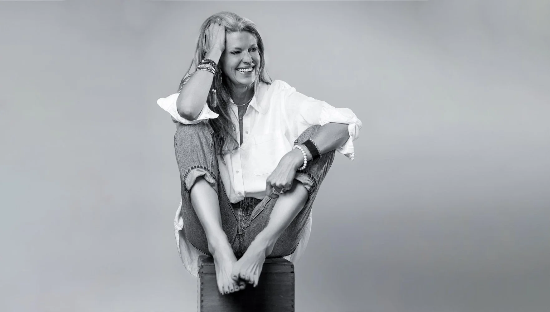 A woman sitting on a wooden box, smiling, with her right hand touching her hair, wearing a white shirt and rolled-up jeans, accessorized with bracelets and necklaces, against a plain background.