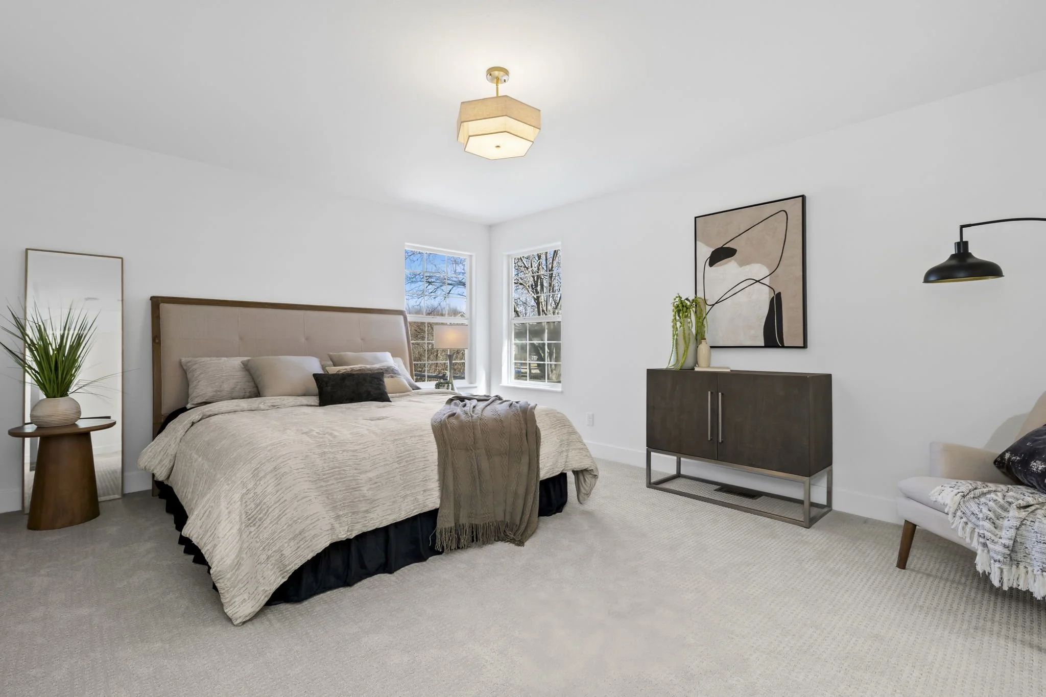 Bright and modern bedroom with a large bed, decorative pillows, and a beige throw blanket. Two windows with views of trees outside, a black dresser with a piece of abstract art hanging above, a potted plant, and a white armchair with a throw blanket. Neutral color palette with beige, white, black, and natural wood accents.