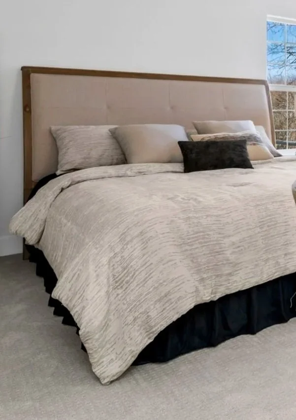 A neatly made bed with a beige comforter, multiple pillows, and a wooden headboard in a bedroom with a window showing an outdoor scene.