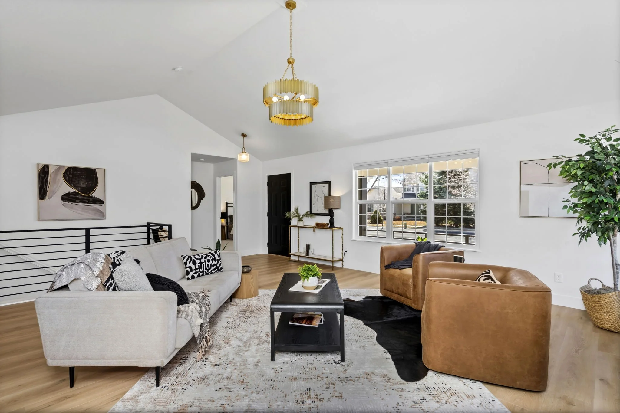 Living room with a white sofa, two tan leather armchairs, a black coffee table, a large window, a white rug, and modern artwork on the walls.