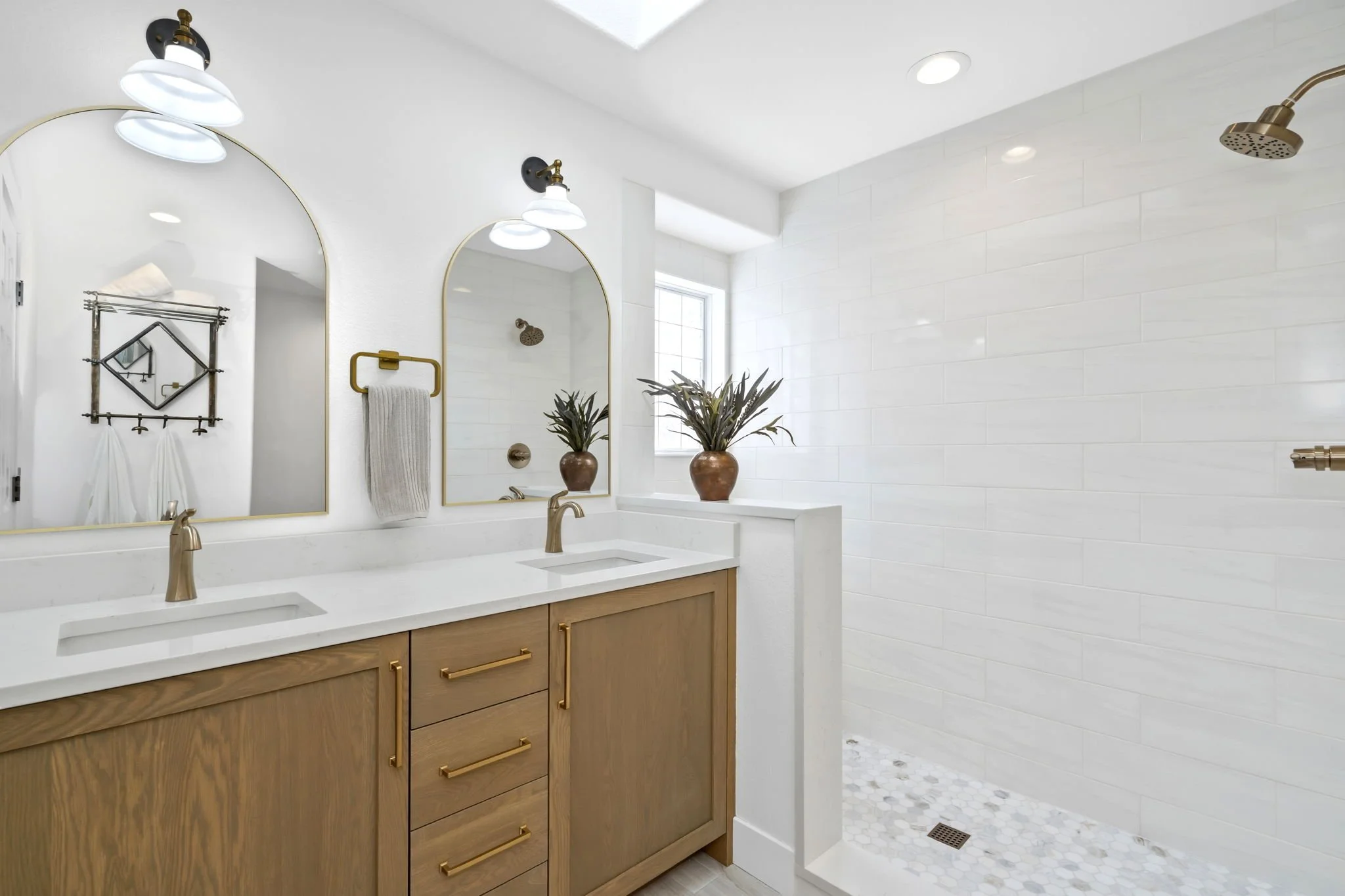 Modern bathroom with double vanity, large mirrors, gold fixtures, white marble countertop, potted plants, and a walk-in shower with white tile walls and a gold showerhead.