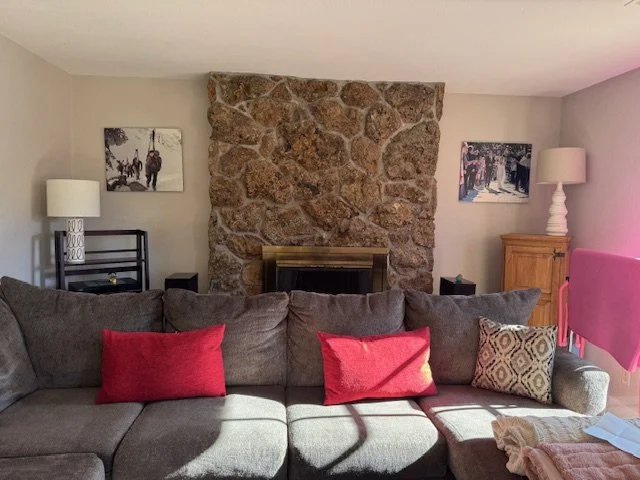 Living room with a gray couch, three red throw pillows, and one patterned pillow in front of a large stone fireplace. Two lamps, one on each side of the fireplace, and framed artwork on the walls.
