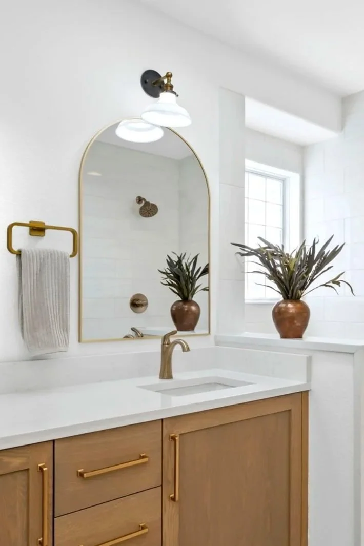A bathroom vanity with a wooden cabinet, a white countertop, and a gold faucet. Above the mirror is a white and bronze light fixture. There is a towel hanging on a gold towel ring, and two potted plants on the windowsill.