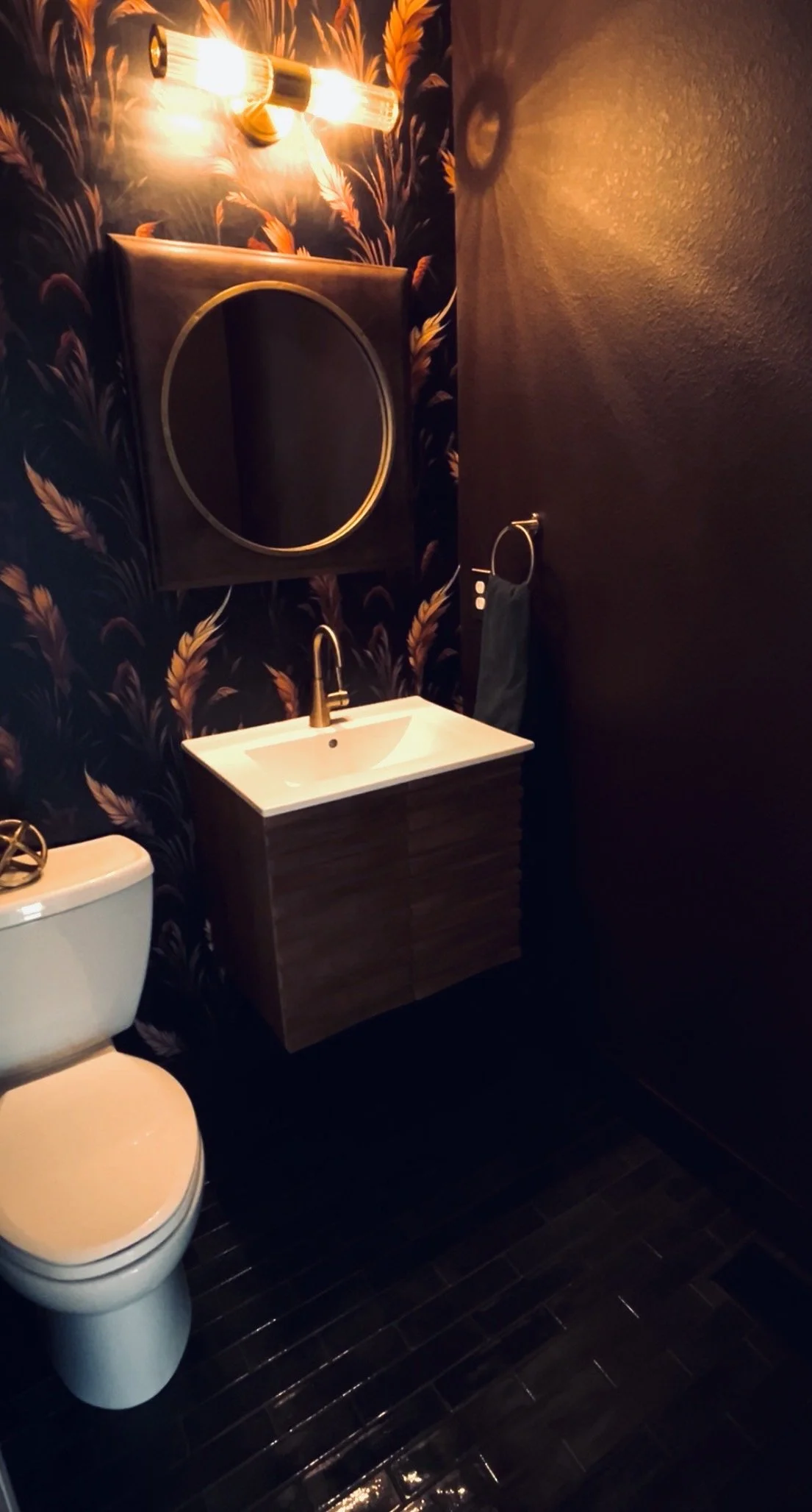 A modern bathroom featuring a toilet, a small wall-mounted sink with a gold faucet, a large circular mirror, decorative wall wallpaper with a feather pattern, and a gold light fixture with exposed bulbs.