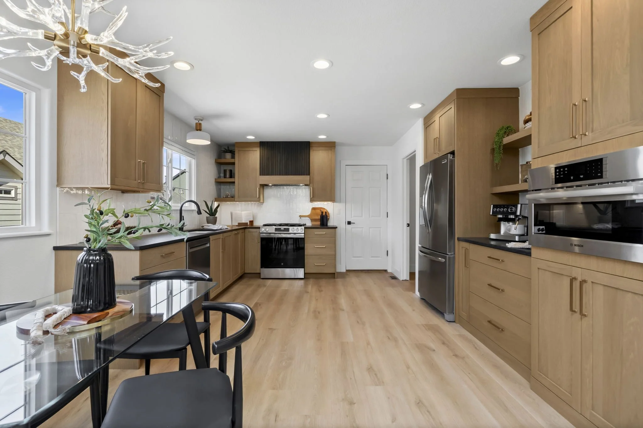 Modern kitchen with light wood cabinets, black countertops, stainless steel appliances, large windows, and hardwood flooring. There is a dining table with black chairs, a decorative vase, and a unique chandelier. The kitchen is bright and spacious.