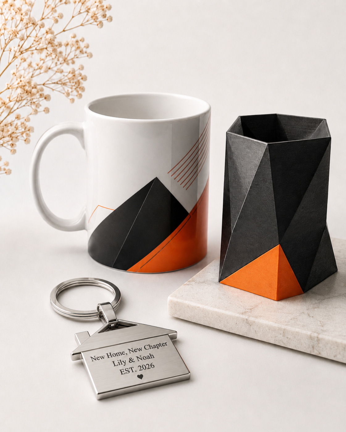 A white ceramic mug and a black geometric vase with orange accents, a metal keychain with engraved message, all placed on a white surface with a beige marble tile and dried flowers in the background.