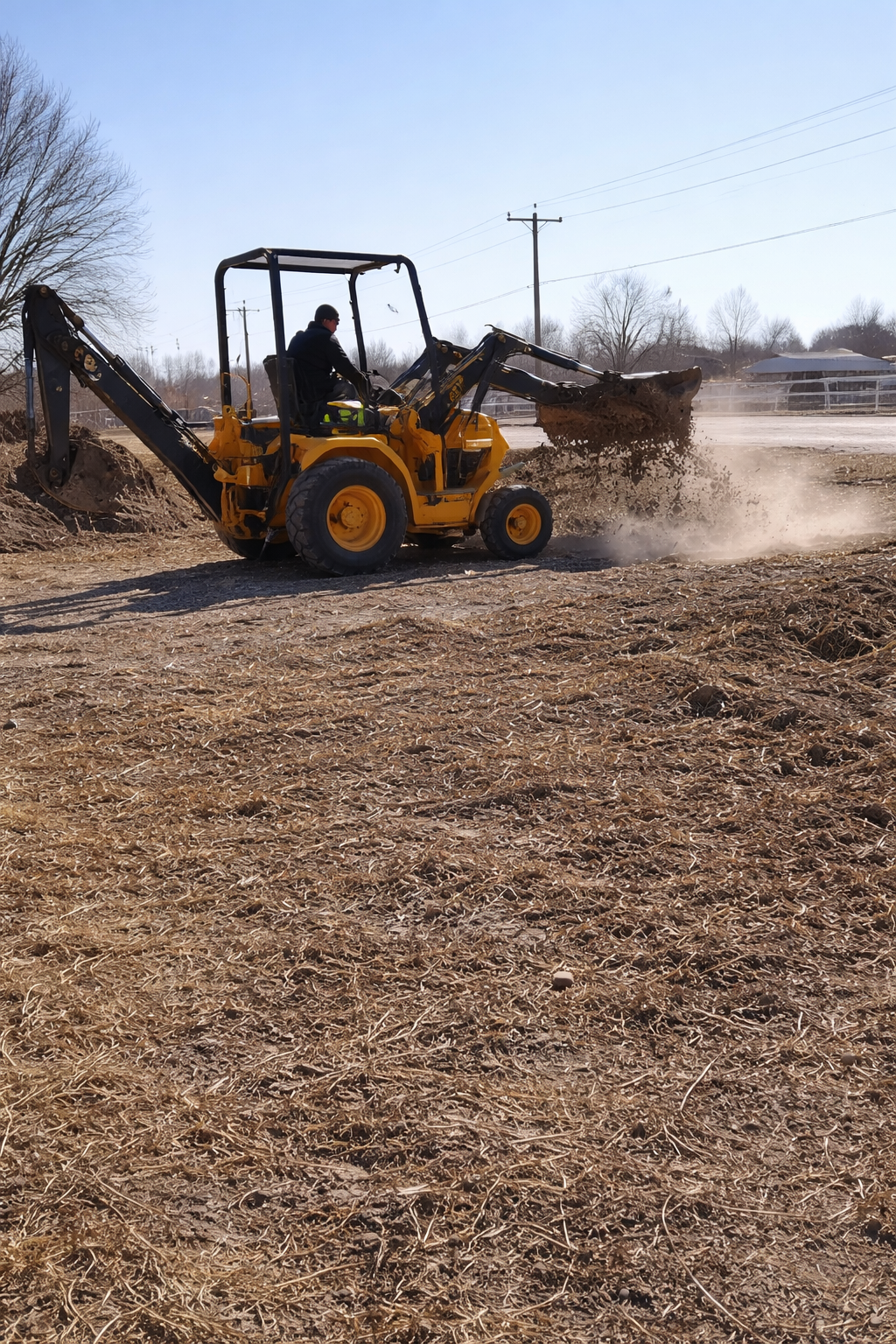 A person operating a yellow backhoe loader digging and moving soil on a dirt field during the daytime.