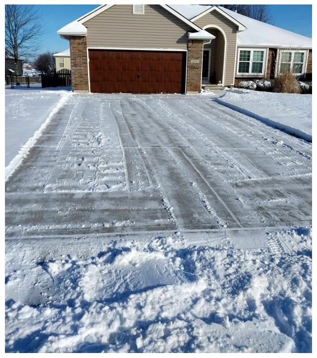 Snow-covered driveway leading to a house with a brown garage door, beige siding, and snow on the roof, with tire tracks visible in the snow.
