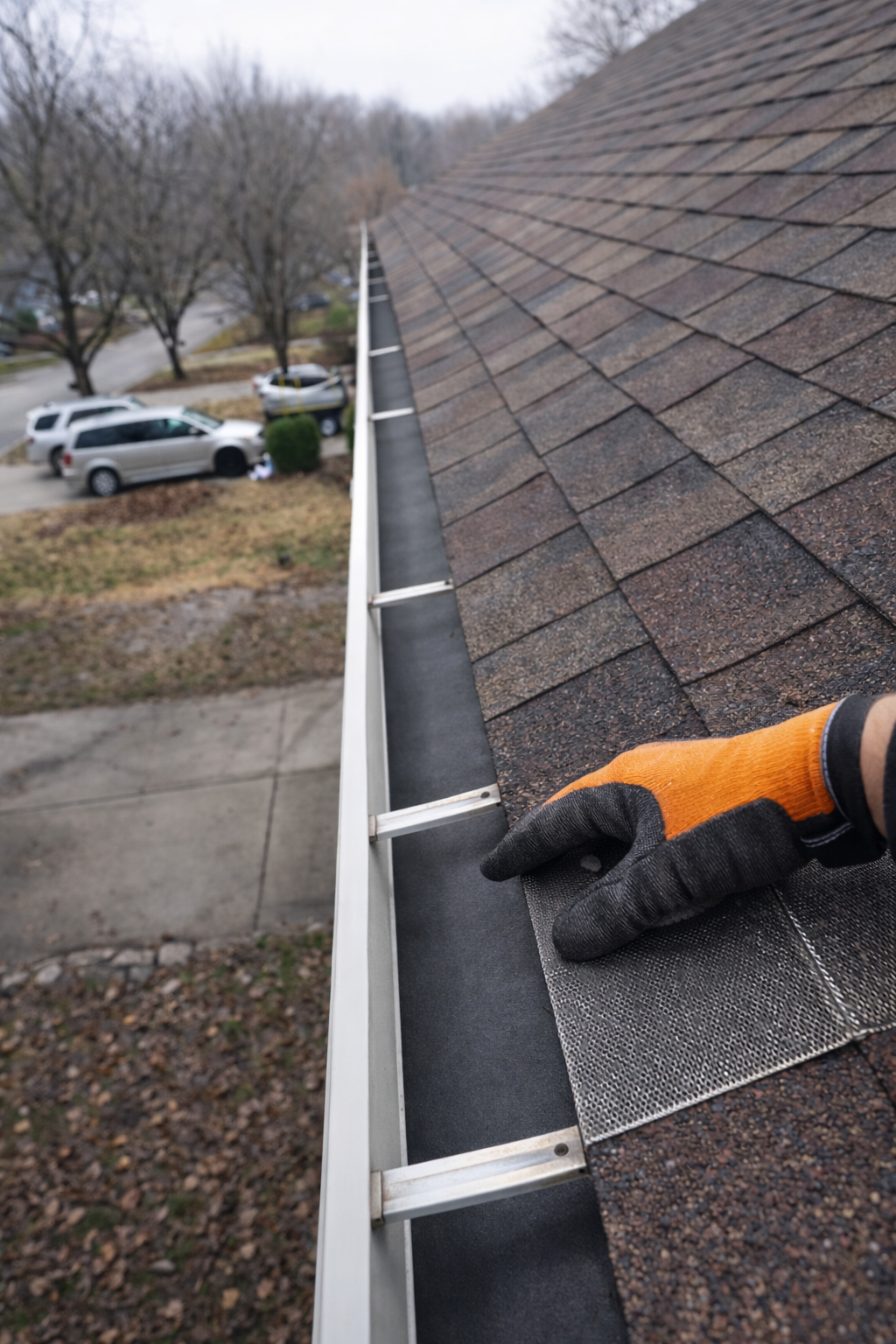 Person installing or repairing a gutter on a sloped shingle roof, with parked cars and leafless trees in the background.
