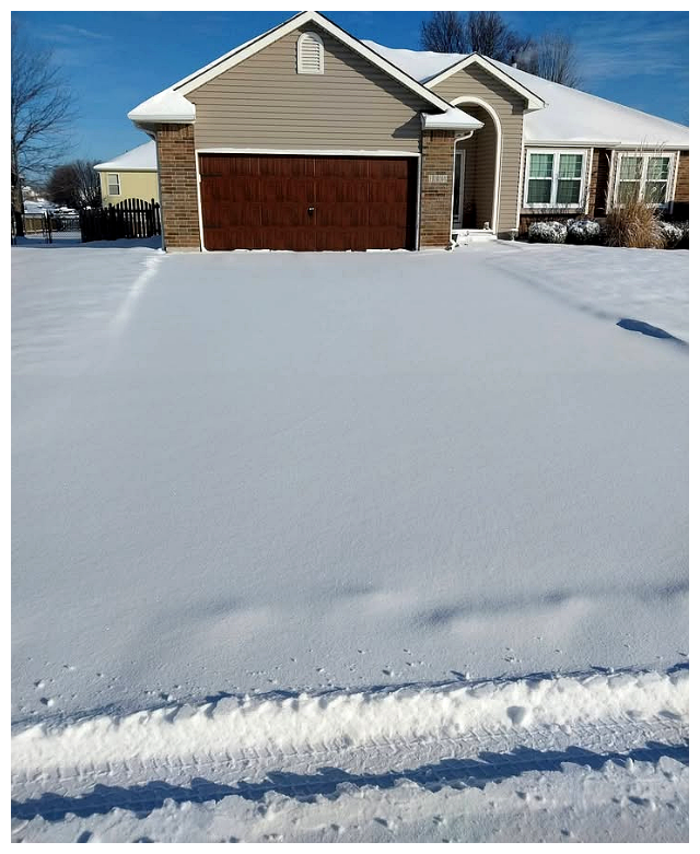 Snow-covered front yard with tire tracks leading to a house with a brown garage door and beige siding.
