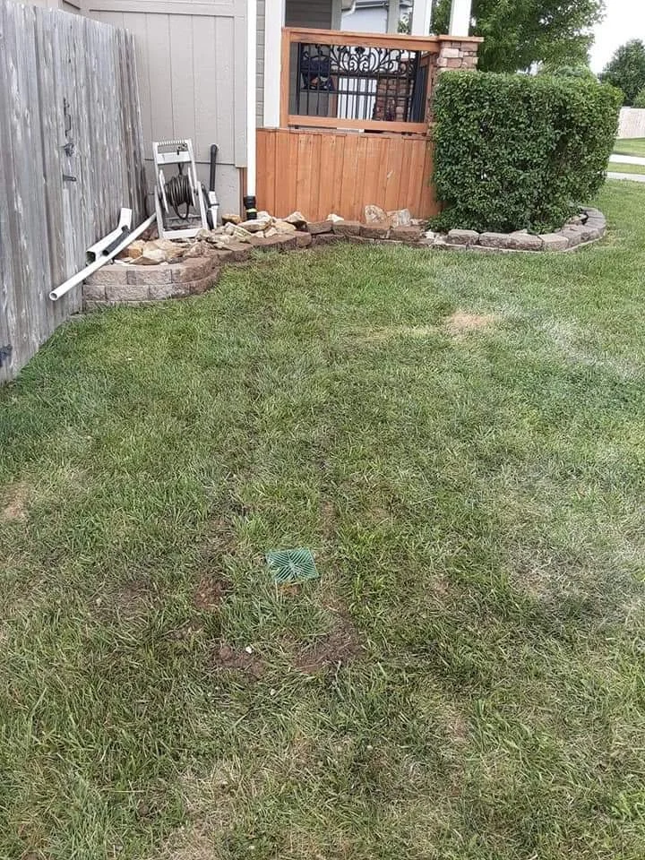 Backyard with a grassy lawn, a corner with solar landscape lights, a wooden privacy fence, a fire hose reel, and a raised porch with a decorative railing, bush, and stone border.