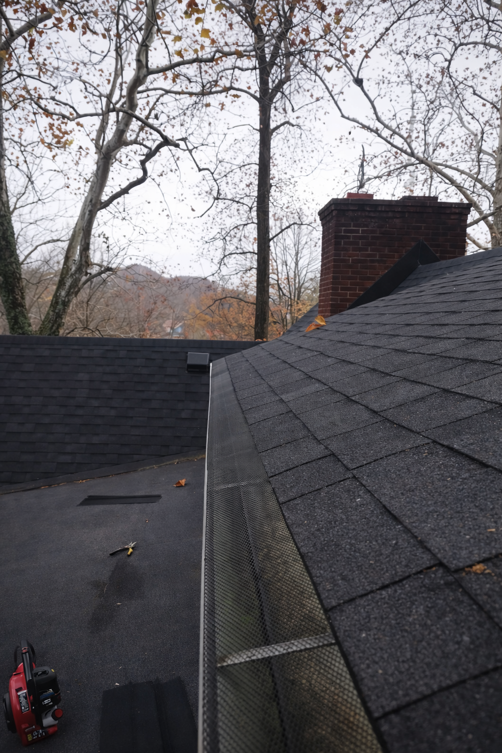 View of a roof with dark asphalt shingles, a brick chimney, and a black vent, with trees and overcast sky in the background.