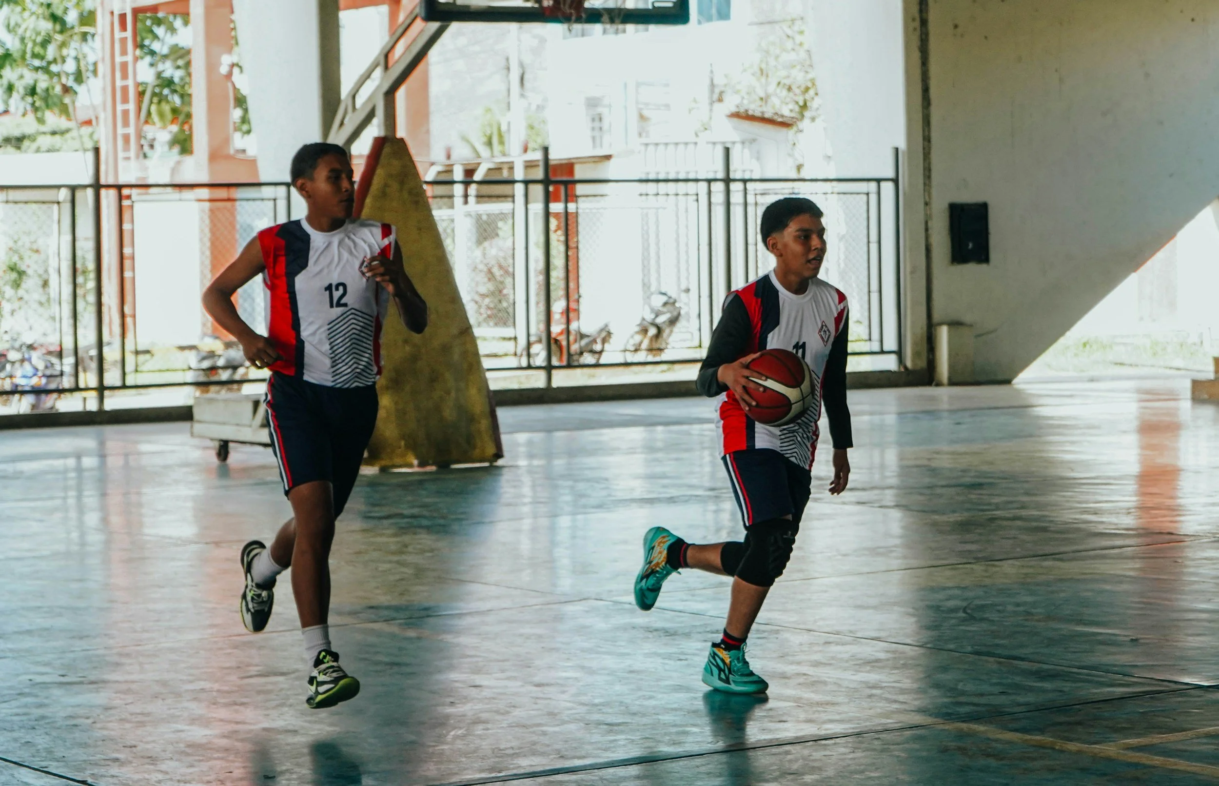 Two young boys playing basketball indoors. One boy is dribbling the ball, while the other is running behind him. They are wearing jerseys and shorts.