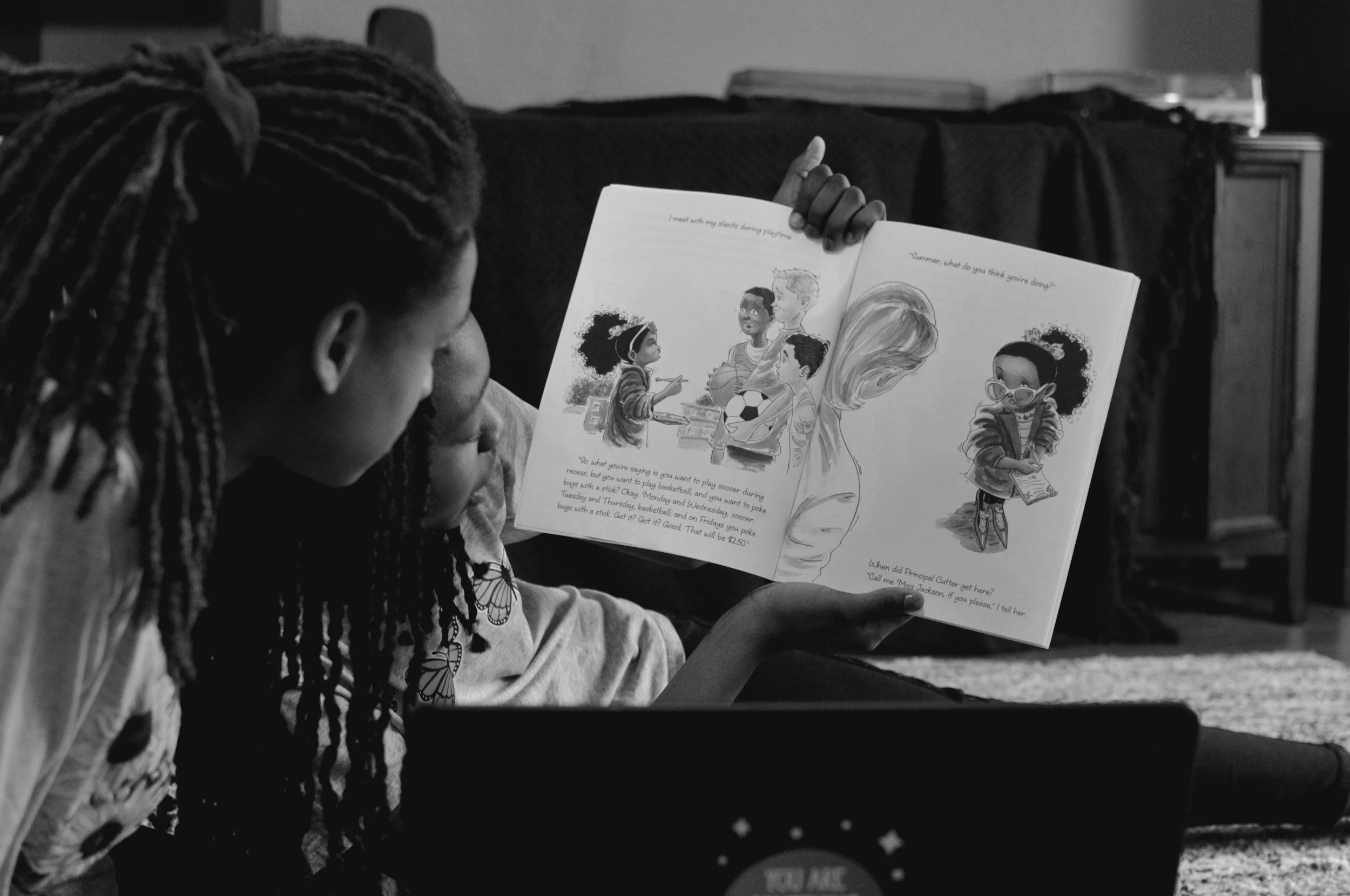 Two girls reading a storybook together on the floor in a living room, with a sofa and scattered books in the background.