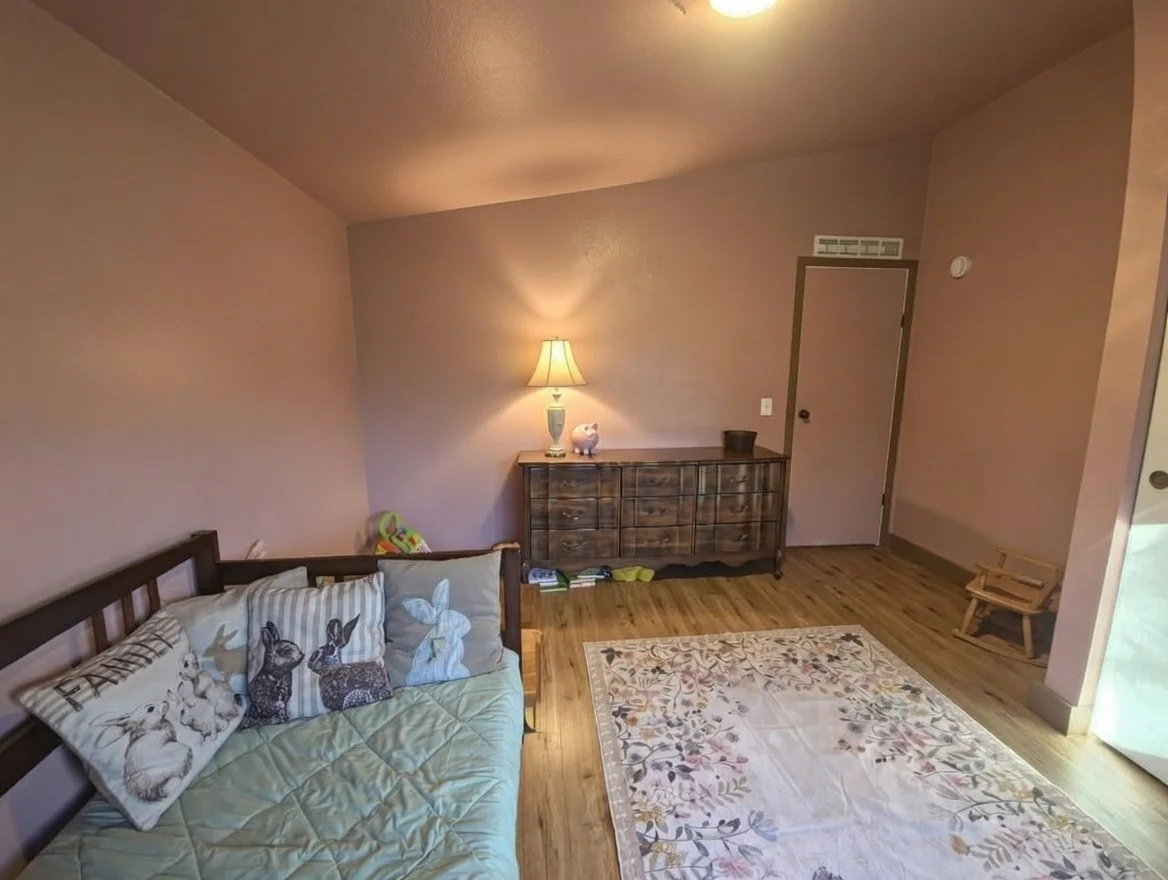 A cozy kids room with a wooden floor, pink walls, a small wooden bench, a white area rug with floral patterns, a wooden sideboard with a lamp, a pig-shaped figurine, and a sofa with bunny-themed pillows.