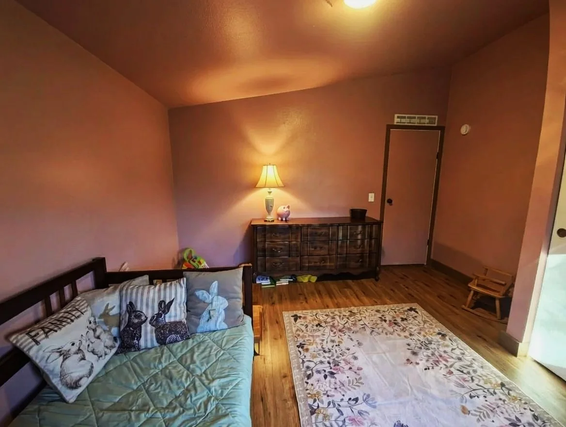 A cozy kids room with a wooden floor, pink walls, a small wooden bench, a white area rug with floral patterns, a wooden sideboard with a lamp, a pig-shaped figurine, and a sofa with bunny-themed pillows.