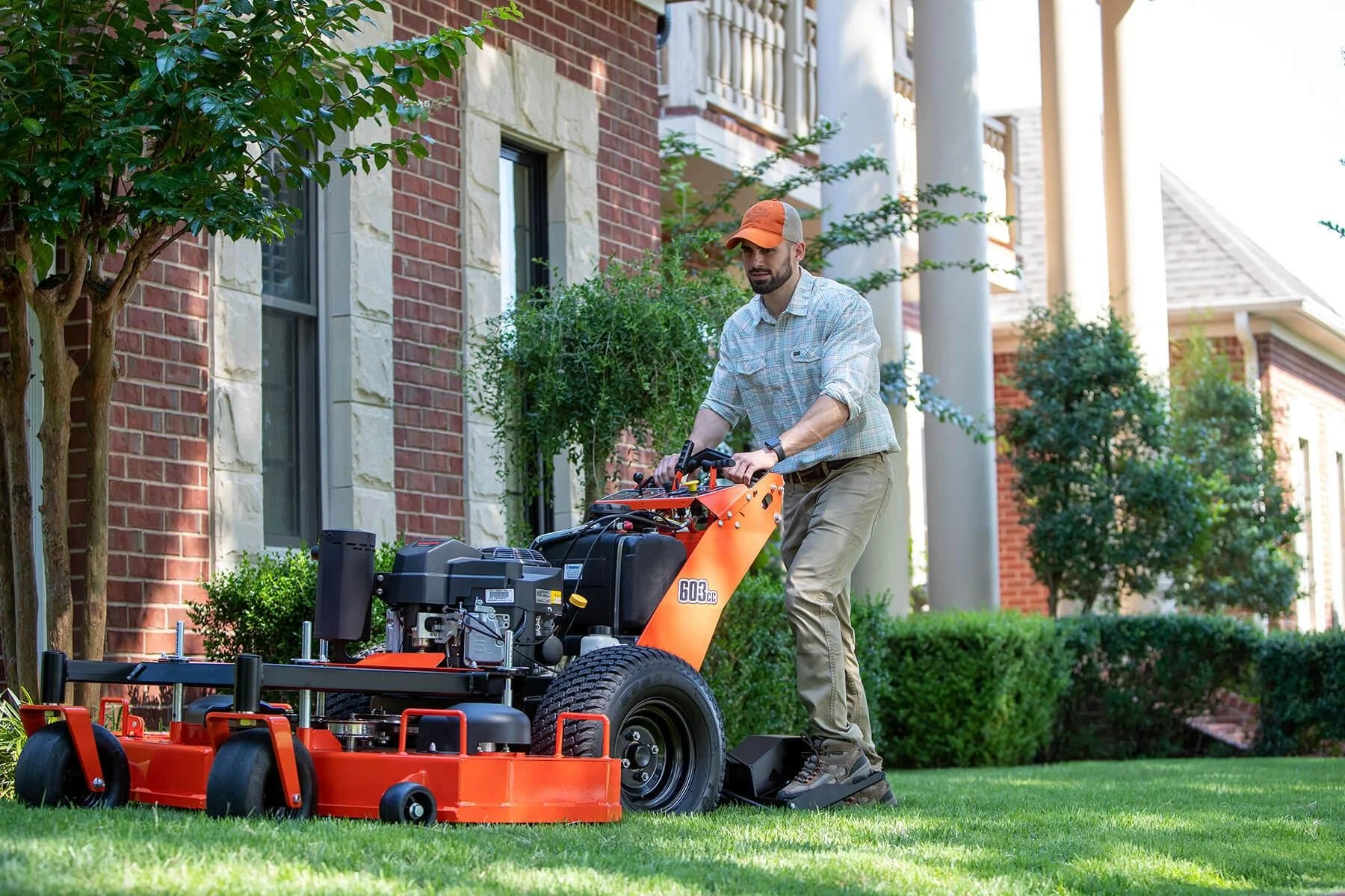 A man operating a riding lawn mower on a well-maintained lawn outside a brick house with white pillars and lush greenery.