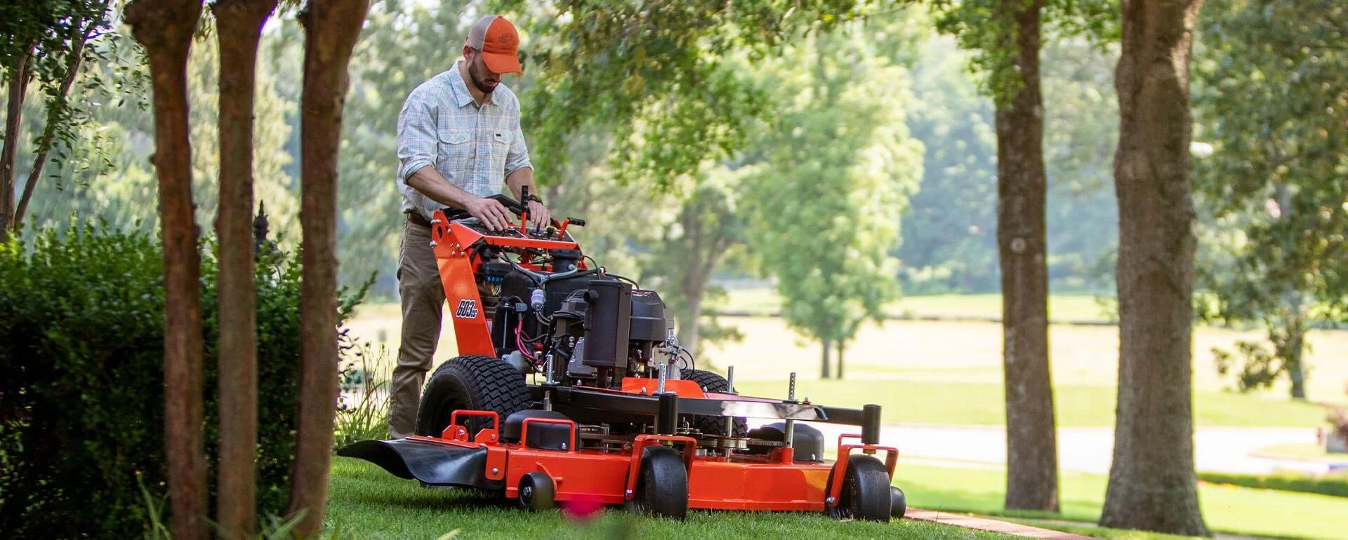 A man operating a large orange mowing machine in a park with green trees and grass.