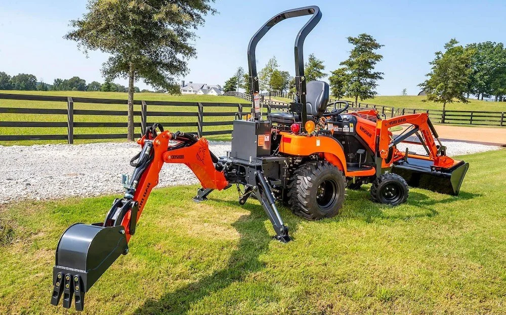 Orange compact utility tractor with a front bucket and rear backhoe attachment on a grassy yard, with trees and a black fence in the background.