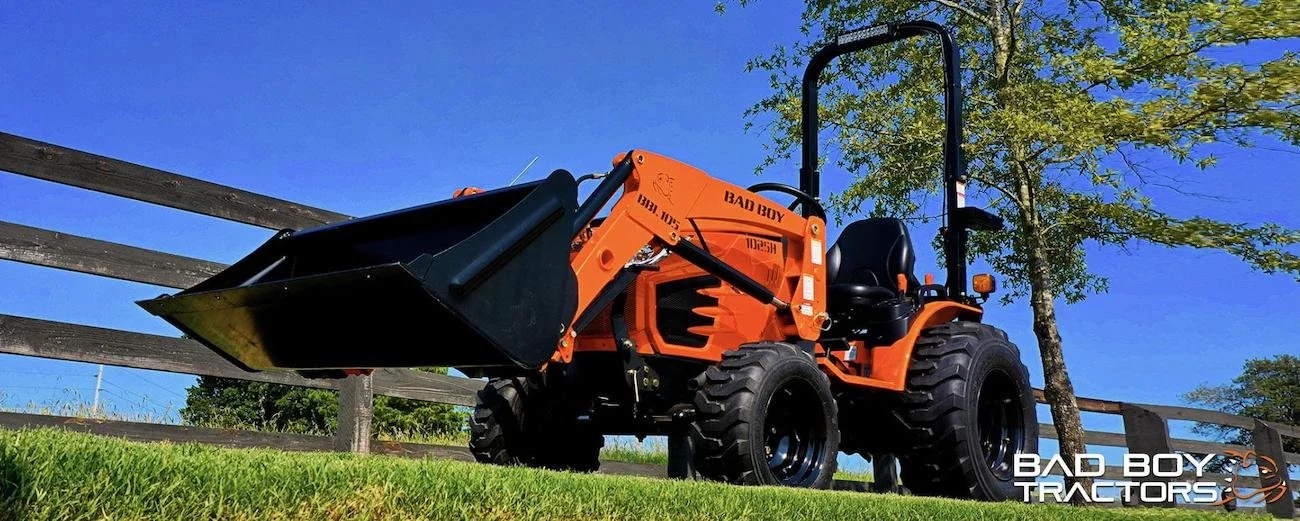 Orange tractor with a front loader attachment parked on grass near a wooden fence with a tree in the background under a clear blue sky, labeled with 'BAD BOY TRACTORS' in the bottom right corner.