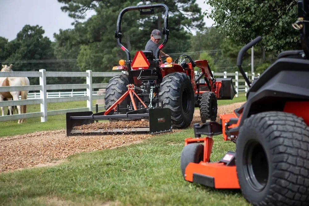 A person operating a red tractor with a blade attachment on a gravel path. Another orange tractor is parked nearby. There is a white fence with horses in the background and trees.