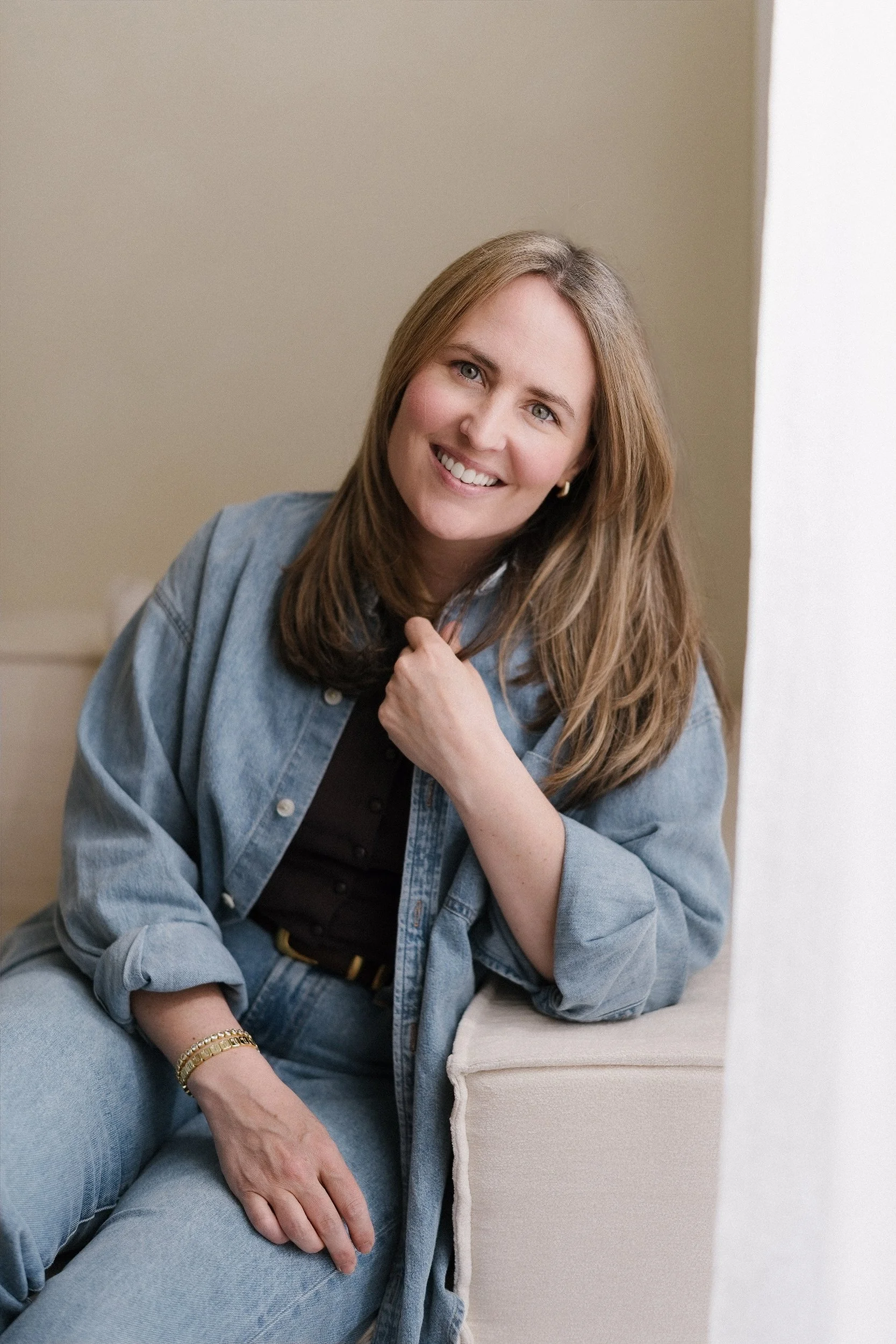 A woman with long brown hair and blue eyes, wearing a denim jacket and black top, sitting on a beige couch, smiling at the camera with her hand on her chest. She is accessorized with gold jewelry.