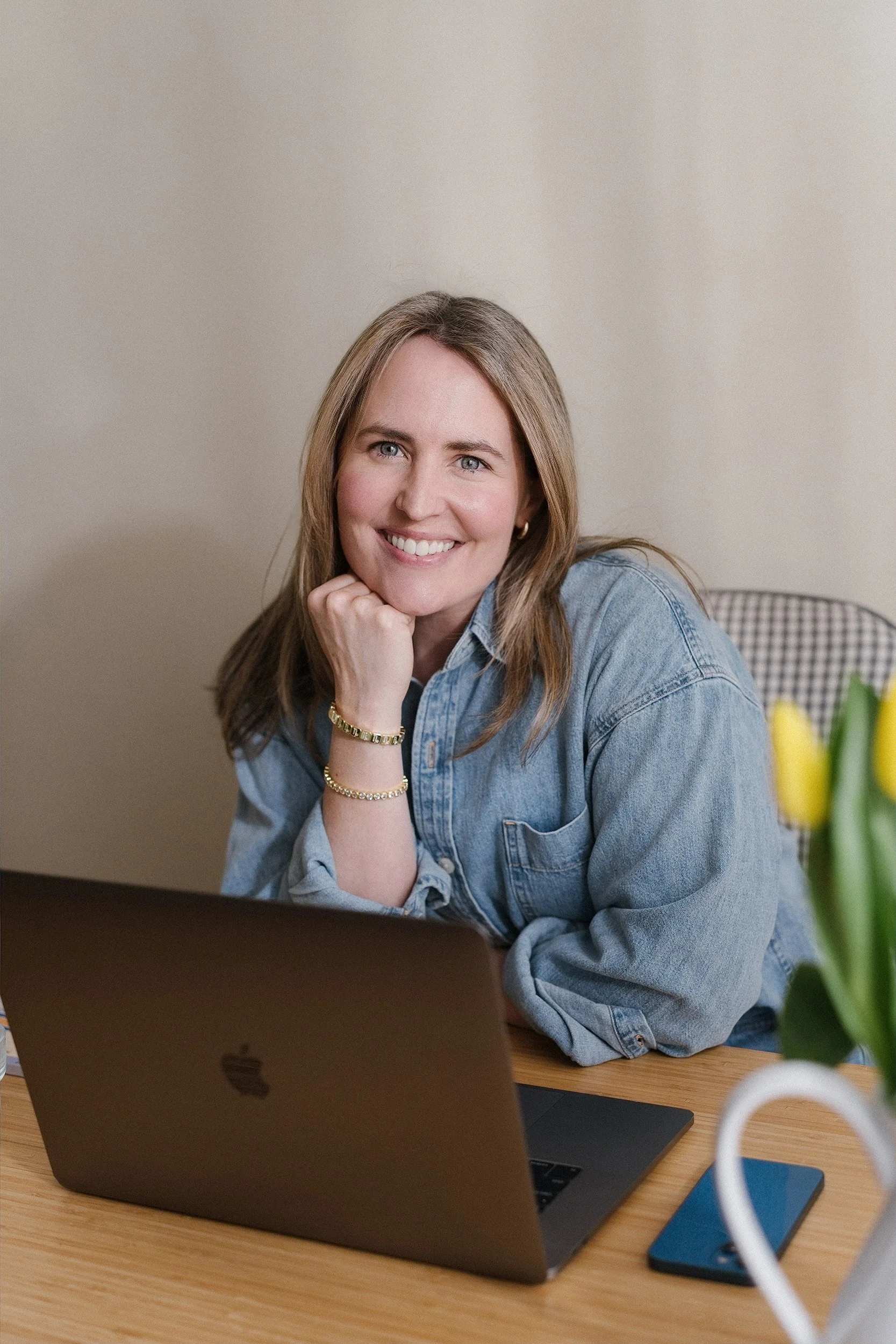 A woman with long blonde hair, wearing a denim shirt, is sitting at a wooden desk with a silver laptop in front of her, smiling and resting her chin on her hand, in a well-lit room.