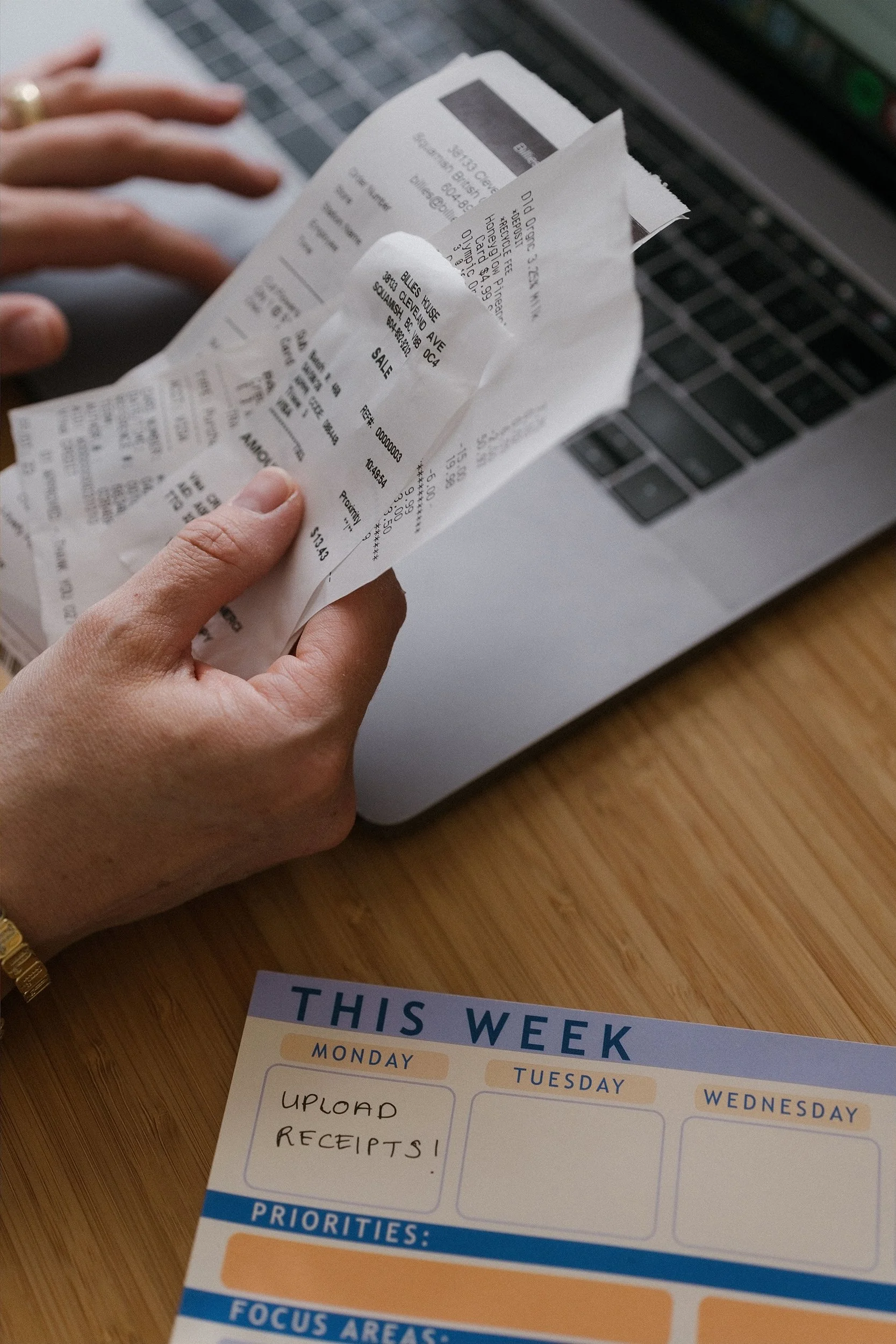 Person holding a receipt above a weekly planner sheet on a wooden desk with a laptop in the background.