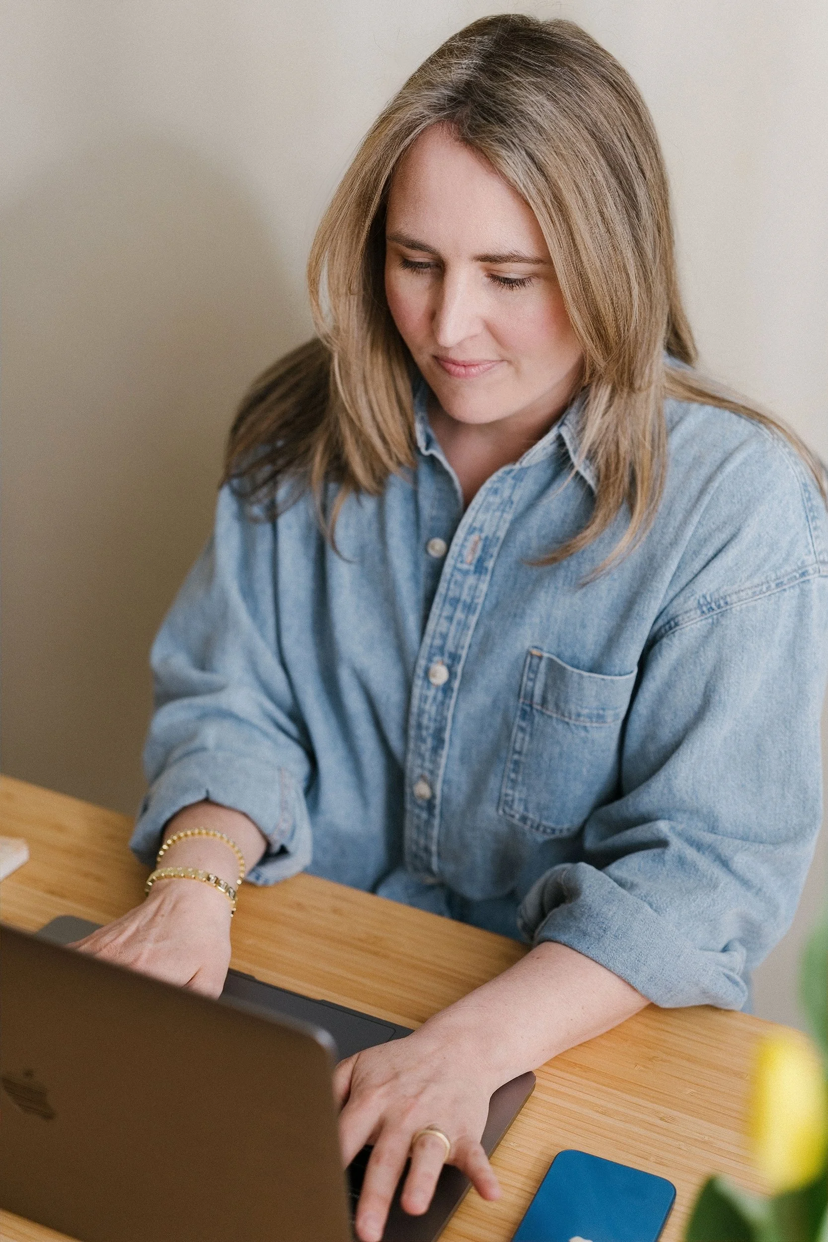 A woman with shoulder-length light brown hair, wearing a denim shirt, working on a laptop at a wooden desk. She has a gold bracelet and ring on her left hand, and her eyes are closed. There is a yellow flower and a blue object on the desk.