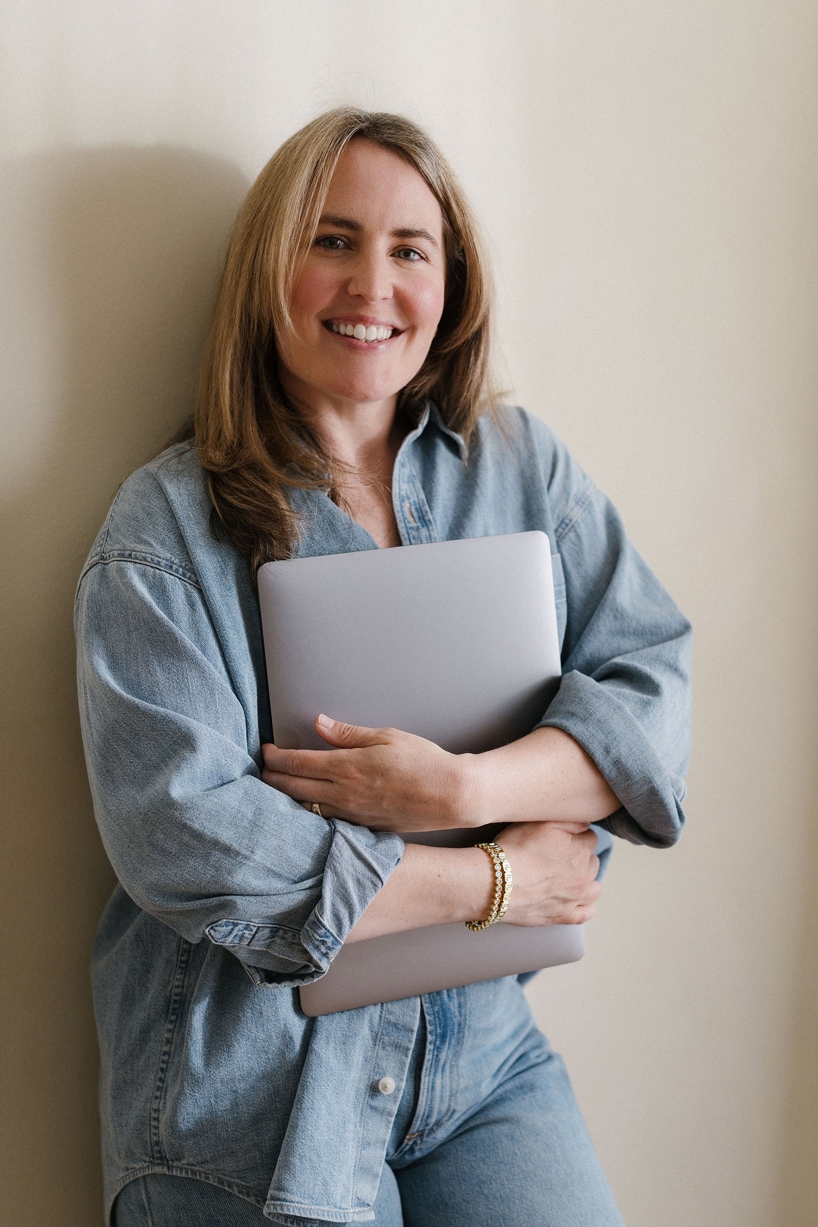 A smiling woman with shoulder-length brown hair wearing a denim shirt and jeans, holding a laptop, standing against a beige wall.