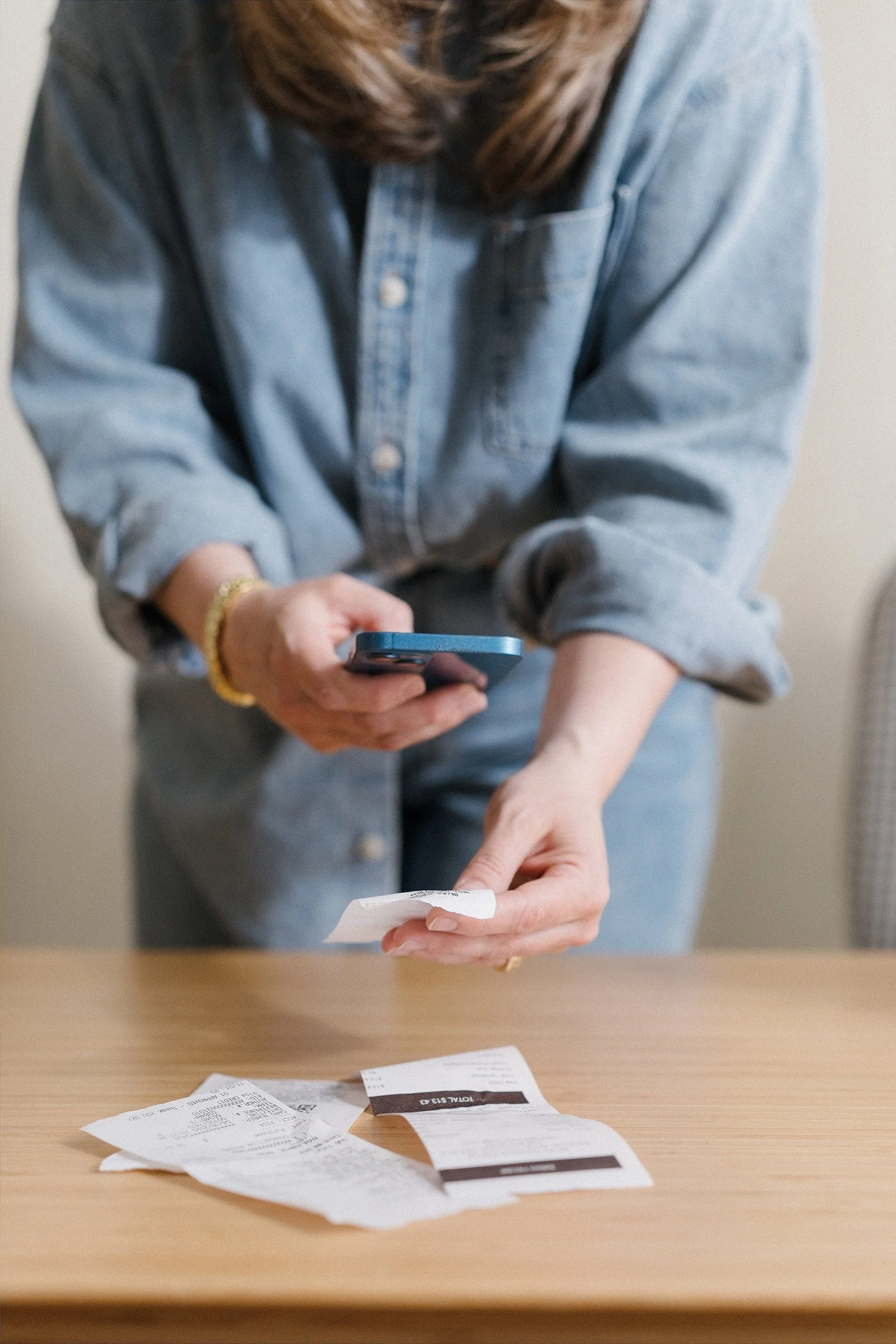 A woman in a blue denim shirt is tearing a receipt with several other receipts on a wooden table. She is holding a blue phone in one hand and tearing the receipt with the other. The table has scattered receipts and a small piece of paper.