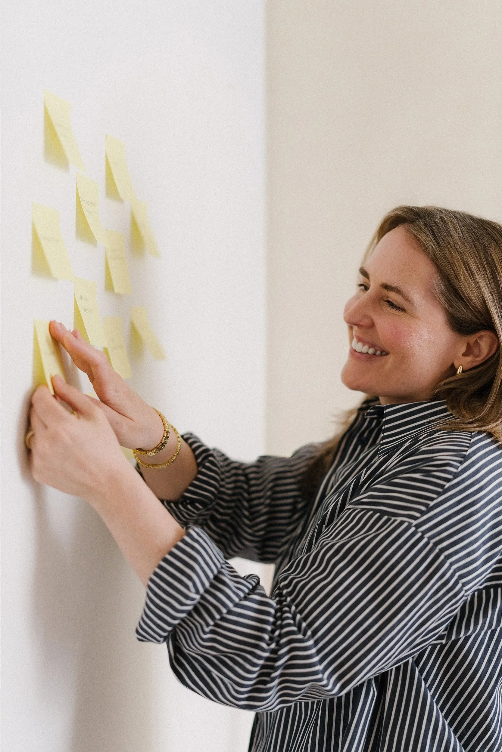 A woman with brown hair, smiling, wearing a black and white striped shirt, is placing sticky notes on a white wall.