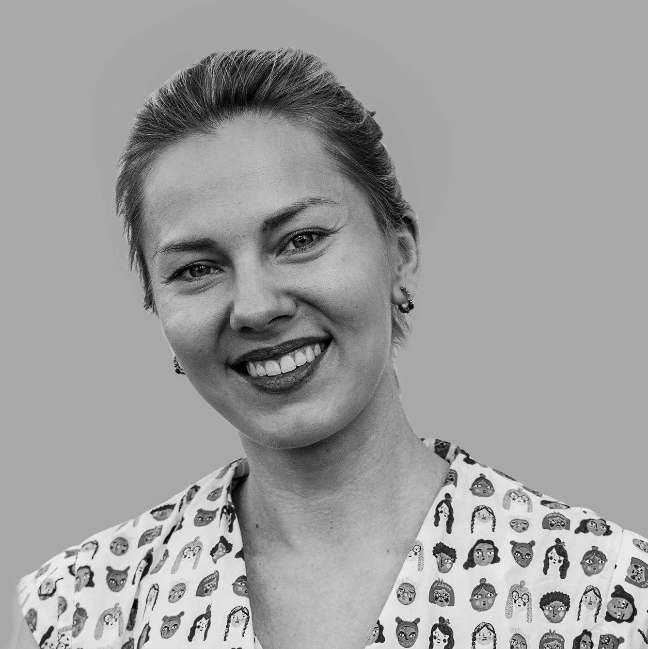 Black and white portrait of a smiling woman with light-colored hair tied back, wearing a shirt with cartoon face illustrations.