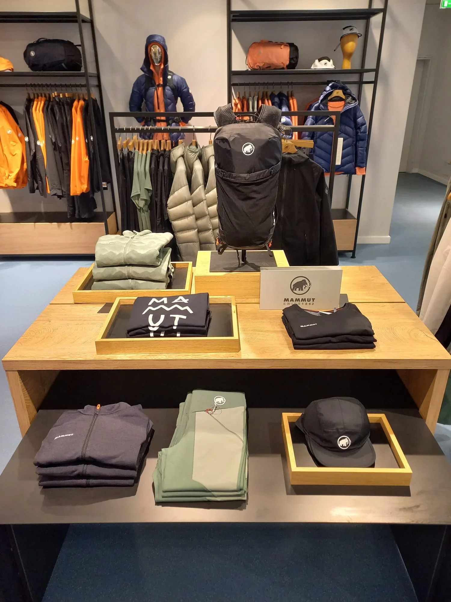 Display of outdoor clothing and gear, including jackets, backpacks, hats, and T-shirts in a retail store, with a mannequin dressed in an outdoor jacket and mask.