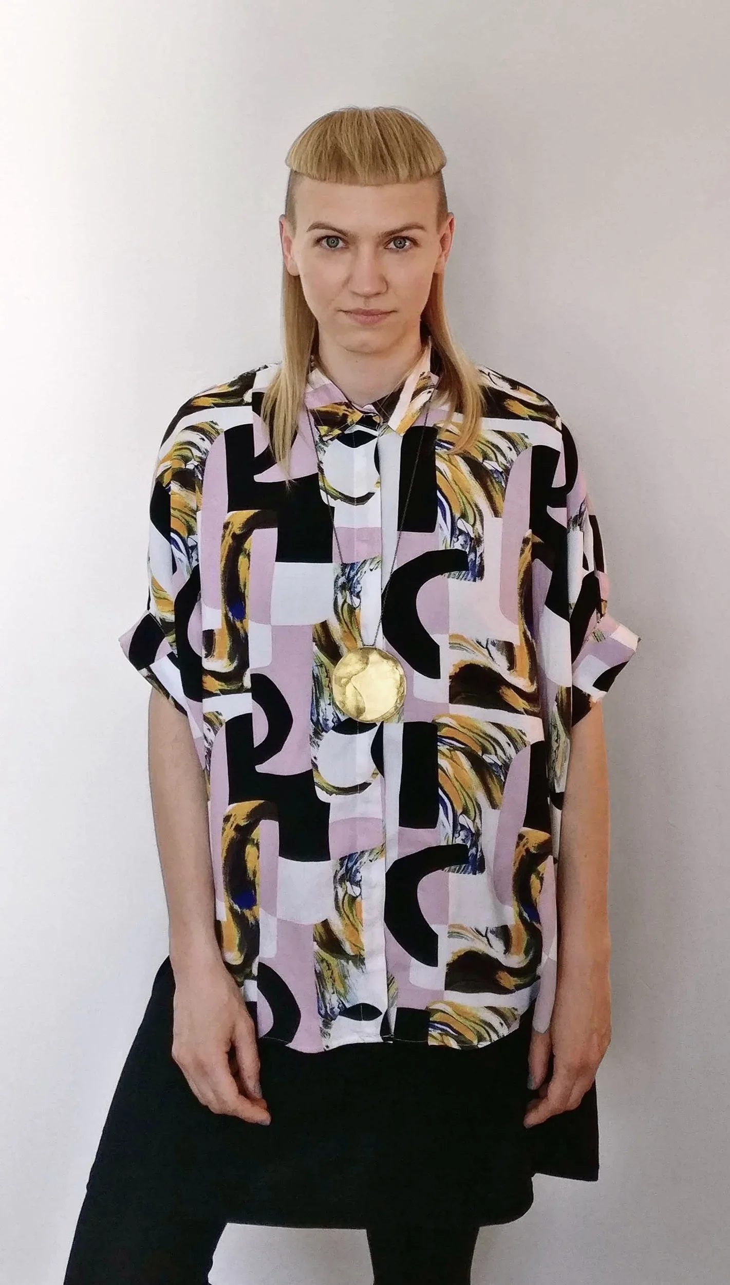A person with Blonde hair in a Madonna style haircut, wearing a colorful abstract patterned shirt with a large gold medal around their neck, standing against a plain white background.