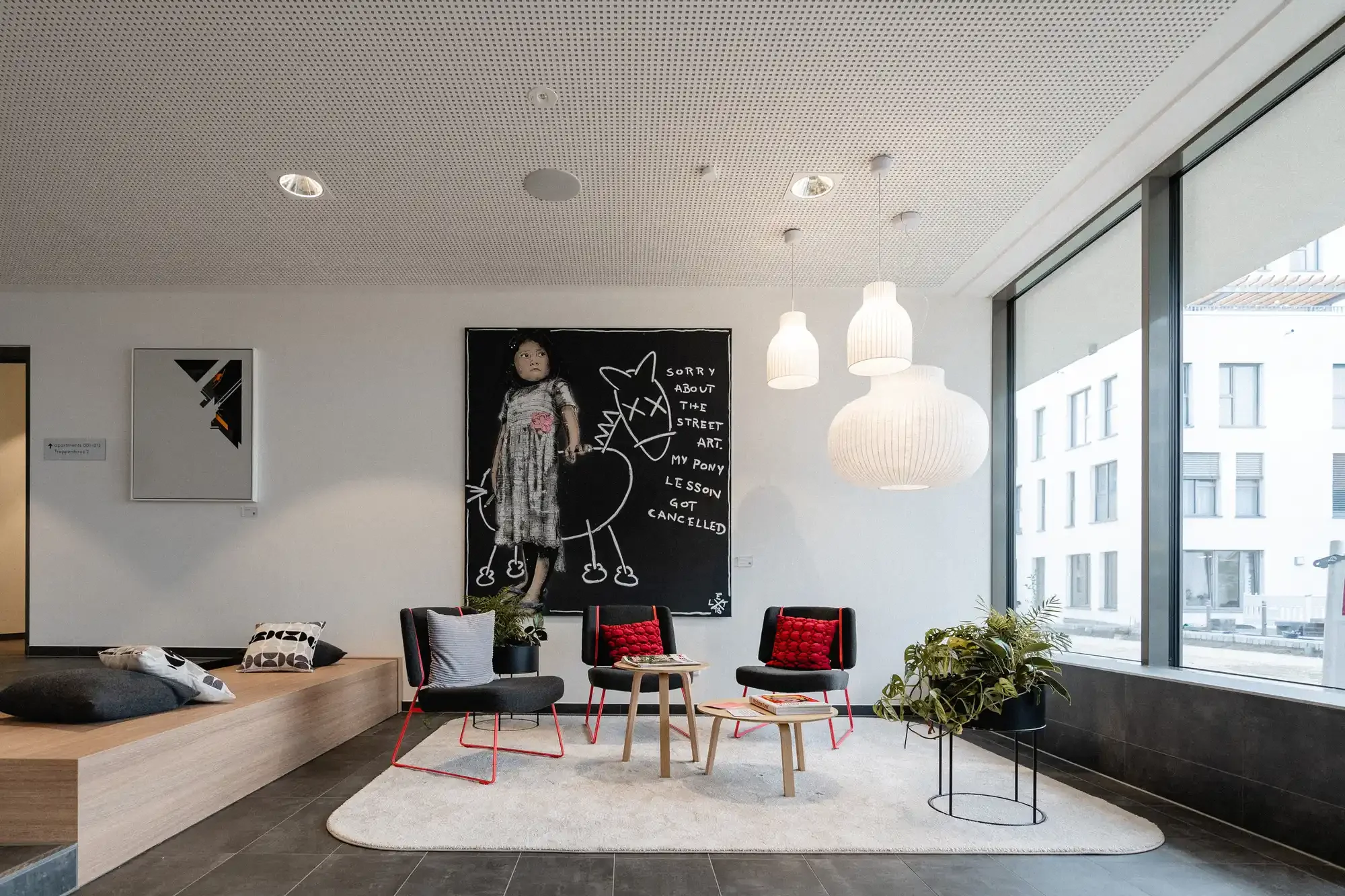 Modern lobby with large windows, black chairs with red pillows, a beige area rug, wooden tables, potted plants, white hanging lamps, and artwork on the walls, including a blackboard with a drawing of a pony and text.