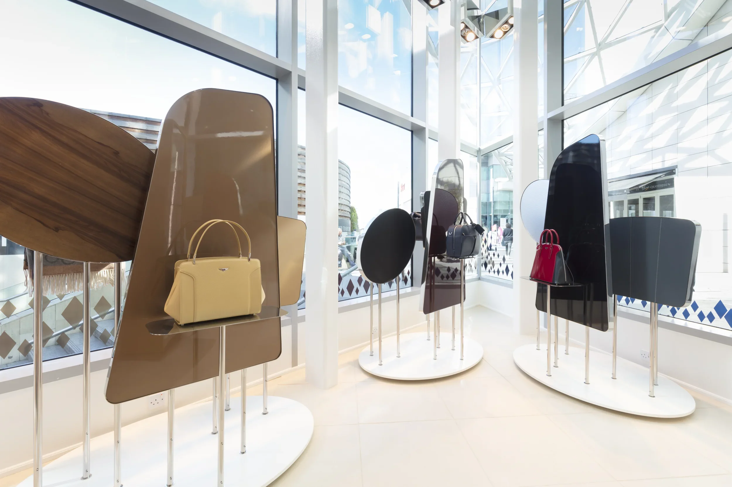 Display of handbags on stands inside a modern, glass-walled store with city view outside.