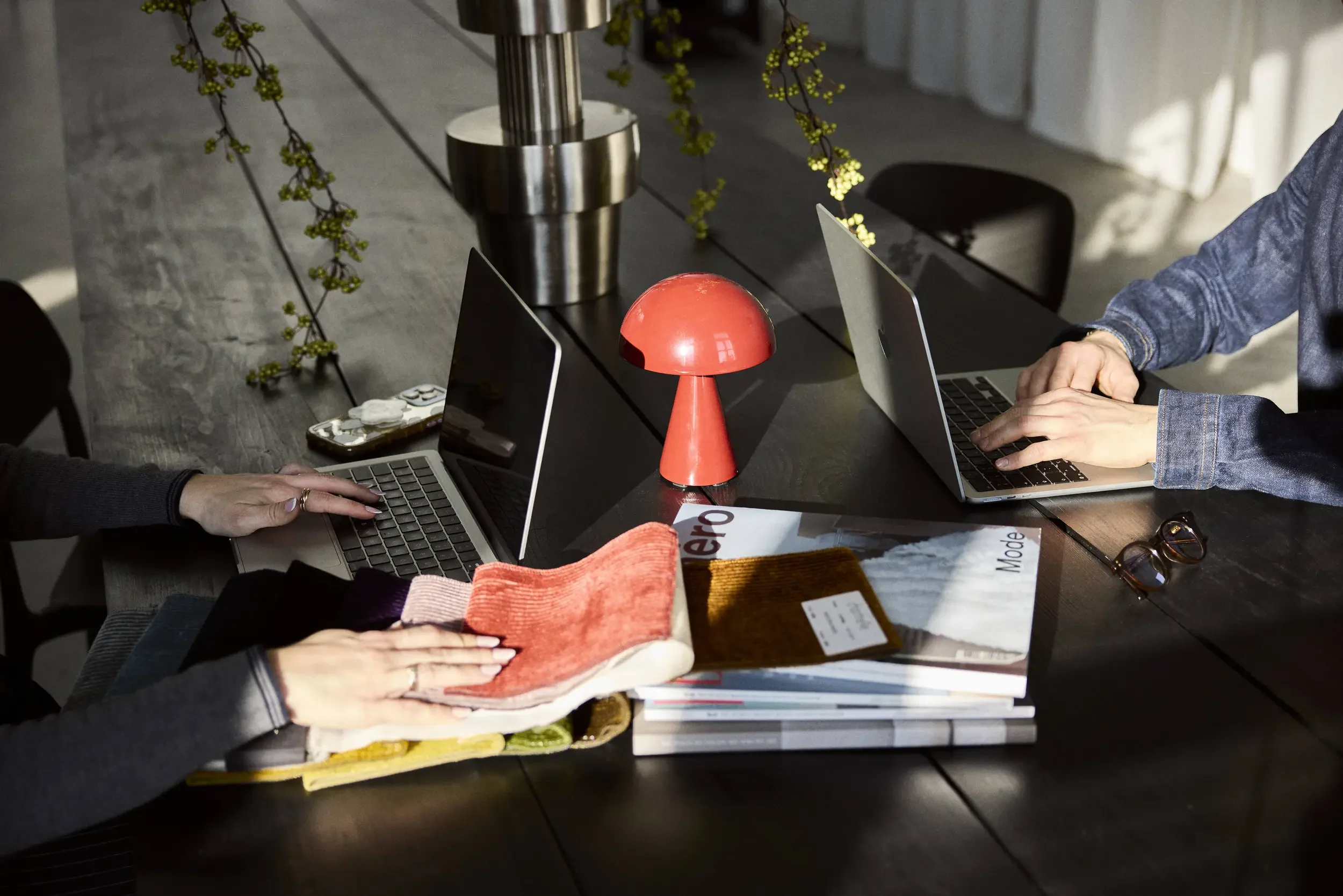 Two people working on laptops at a dark wooden table with fabric samples and magazines, a unique red mushroom-shaped lamp, and small plants in the background.