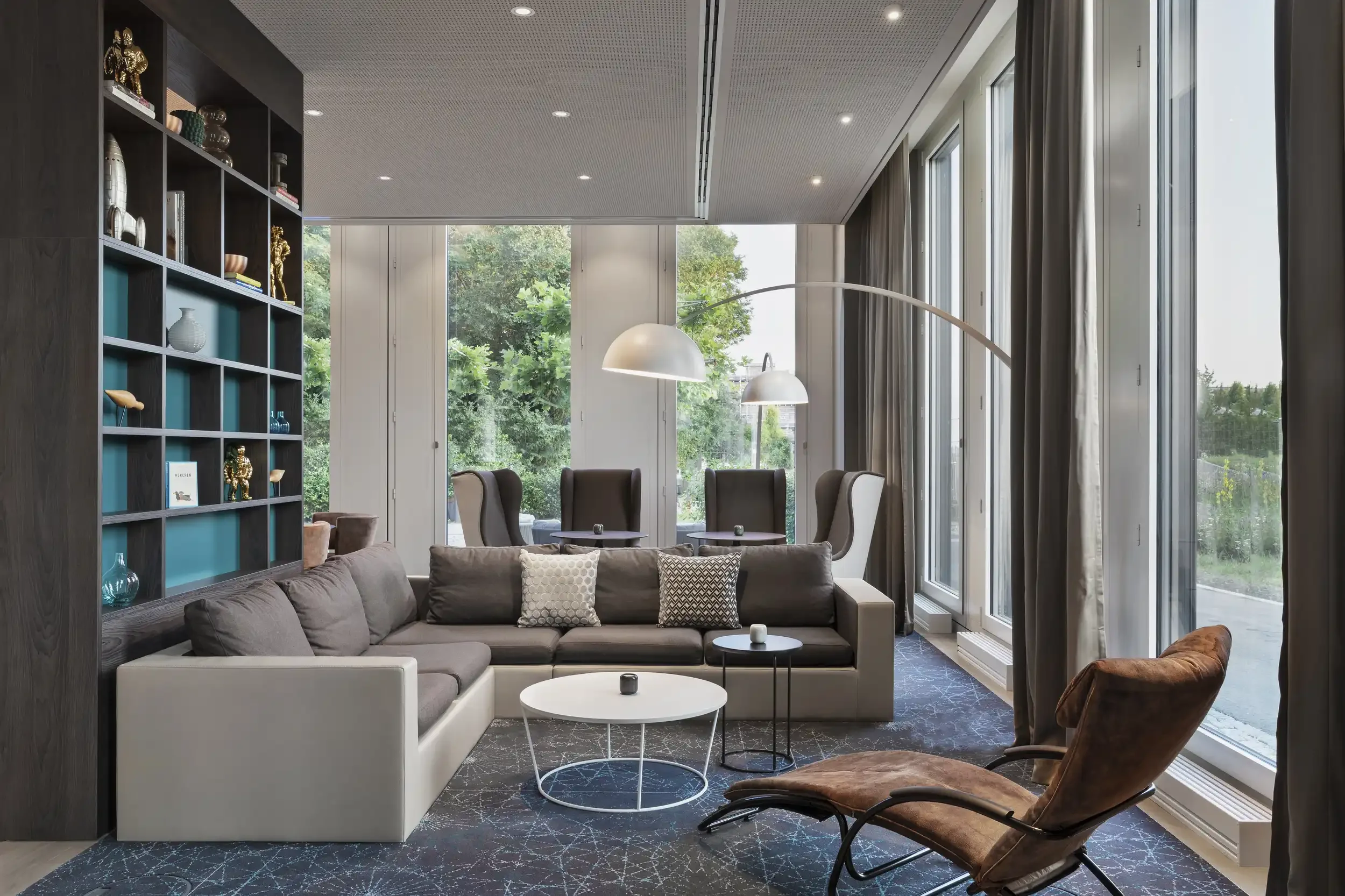 Modern living room with a beige and brown sofa, a brown lounge chair, a bookshelf filled with decorative items, large windows with curtains, and a central white coffee table.