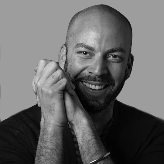 Black and white portrait of a smiling bald man with a beard, resting his chin on his hands, wearing a dark shirt and bracelets on his wrists.