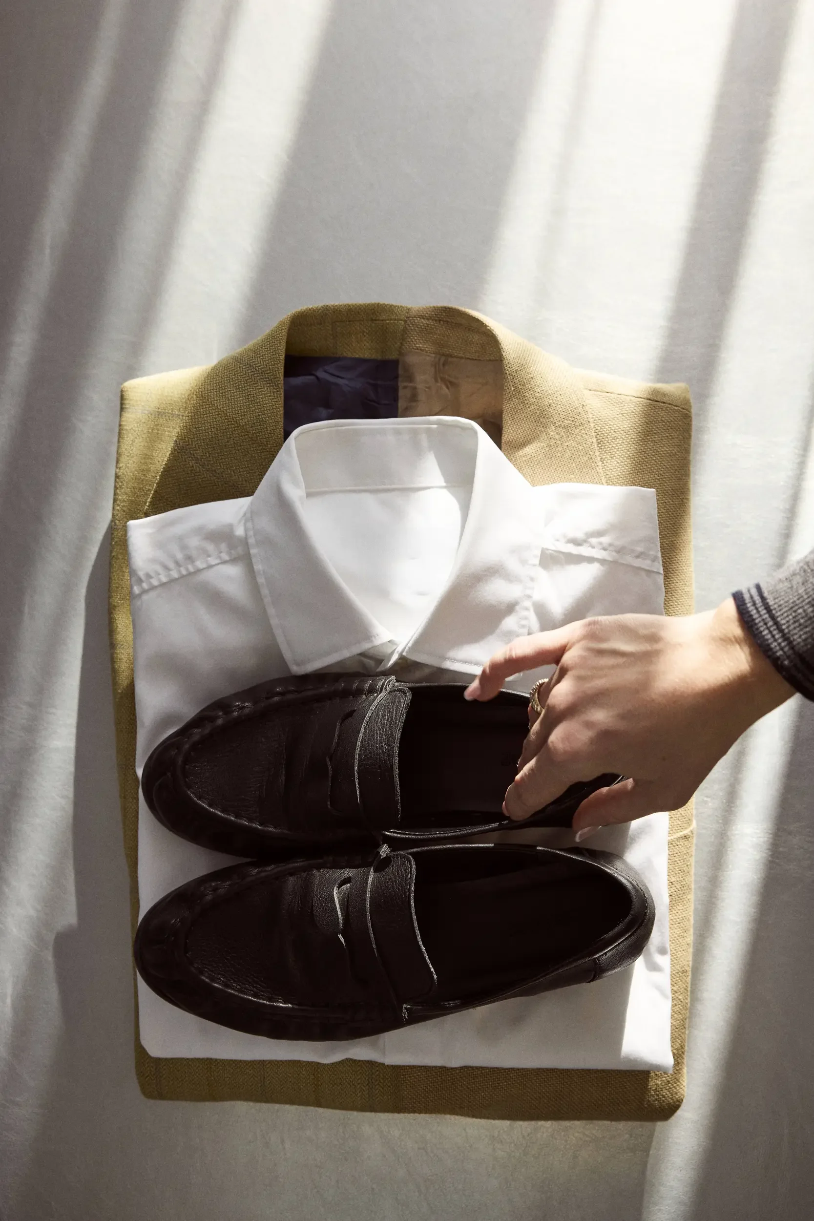 A stack of clothing including a brown blazer, a white dress shirt, and a pair of black shoes on a white shirt, with a hand reaching towards the shoes.