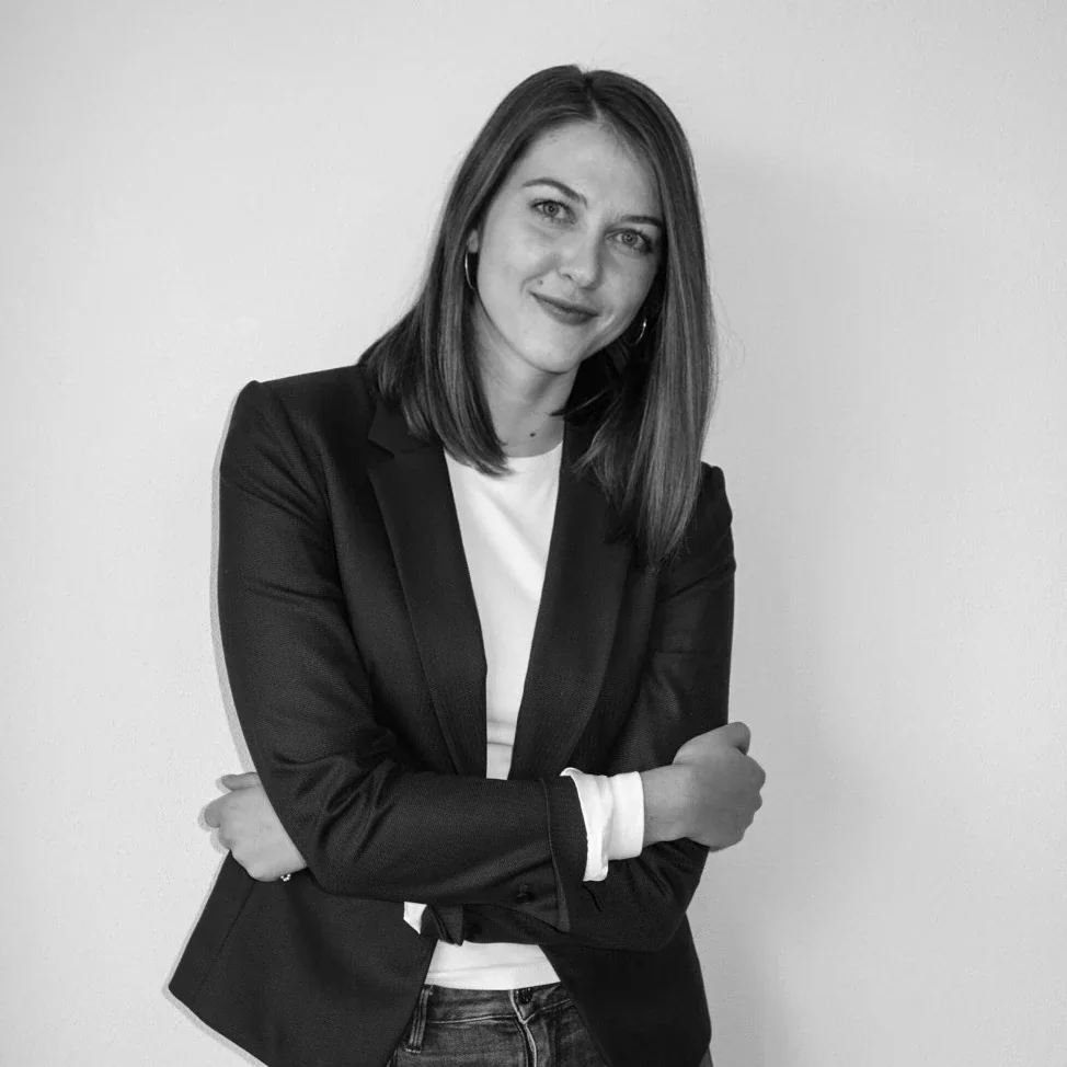 A young woman with shoulder-length brown hair, wearing a black blazer over a white top, smiling while standing against a plain white wall.