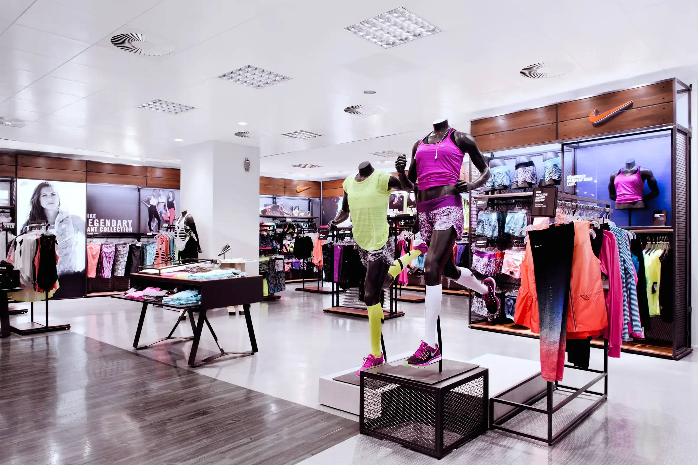 Inside a Nike sporting apparel store, mannequins display colorful athletic outfits, with racks of sportswear and accessories around, and promotional signage in the background.