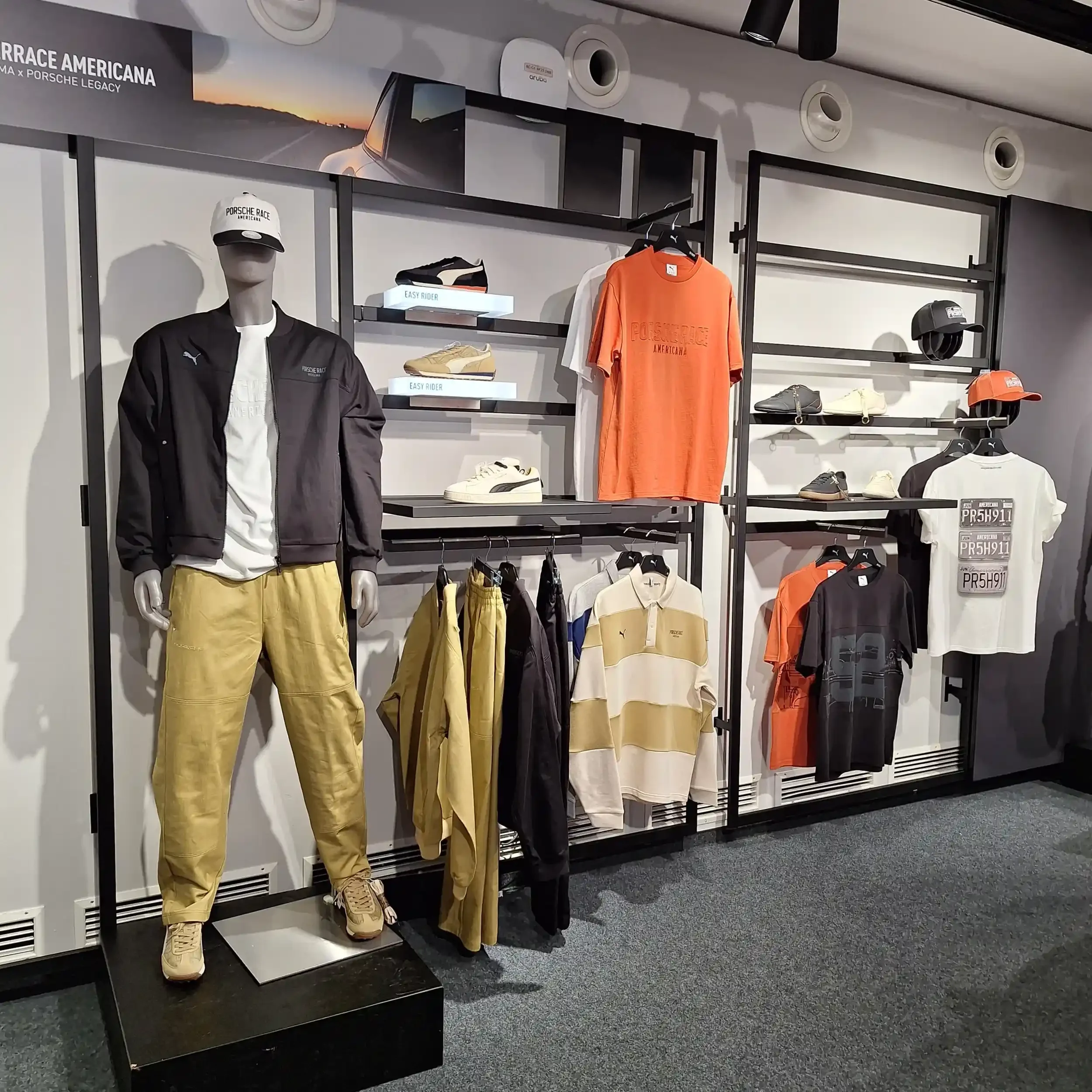 Display of Porsche-themed clothing and accessories including jackets, shirts, pants, and hats in a retail store, with a mannequin wearing a black jacket, white shirt, and yellow pants in the foreground.