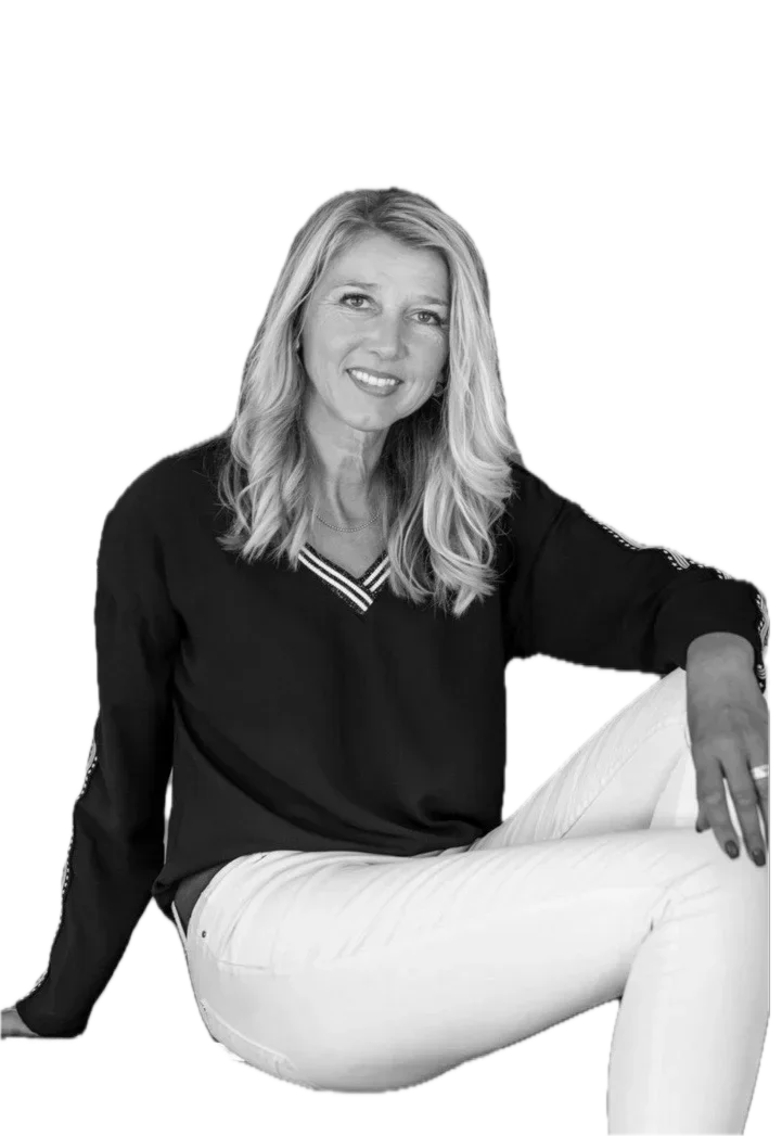 Black and white photo of a woman with long wavy hair, wearing a dark long-sleeve top with a V-neck and white pants, sitting relaxed with one arm resting on her knee, smiling at the camera.
